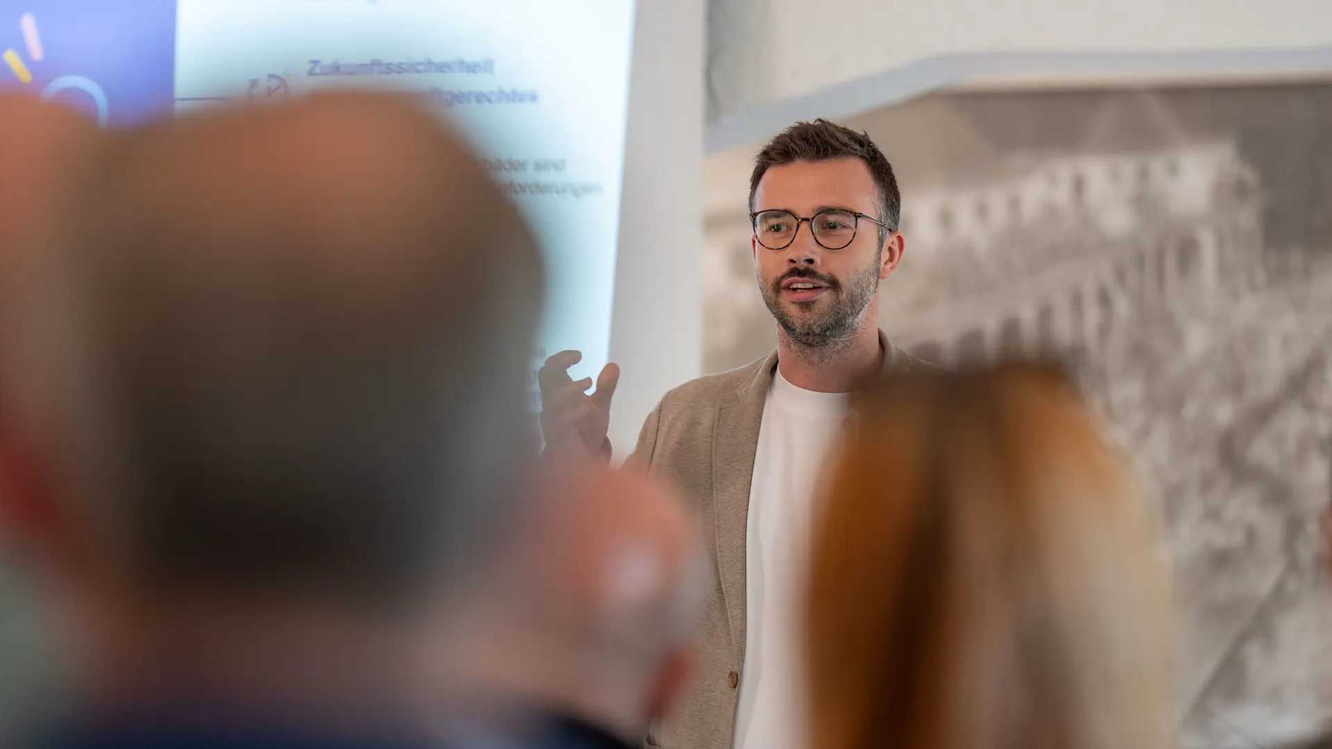 Strategisch handeln, nachhaltig wirken. Eine Person mit Brille und beigem Sakko hält einen Vortrag vor Publikum. Die Zuhörenden sind im Vordergrund unscharf zu sehen, während im Hintergrund eine Präsentationsfolie projiziert wird.