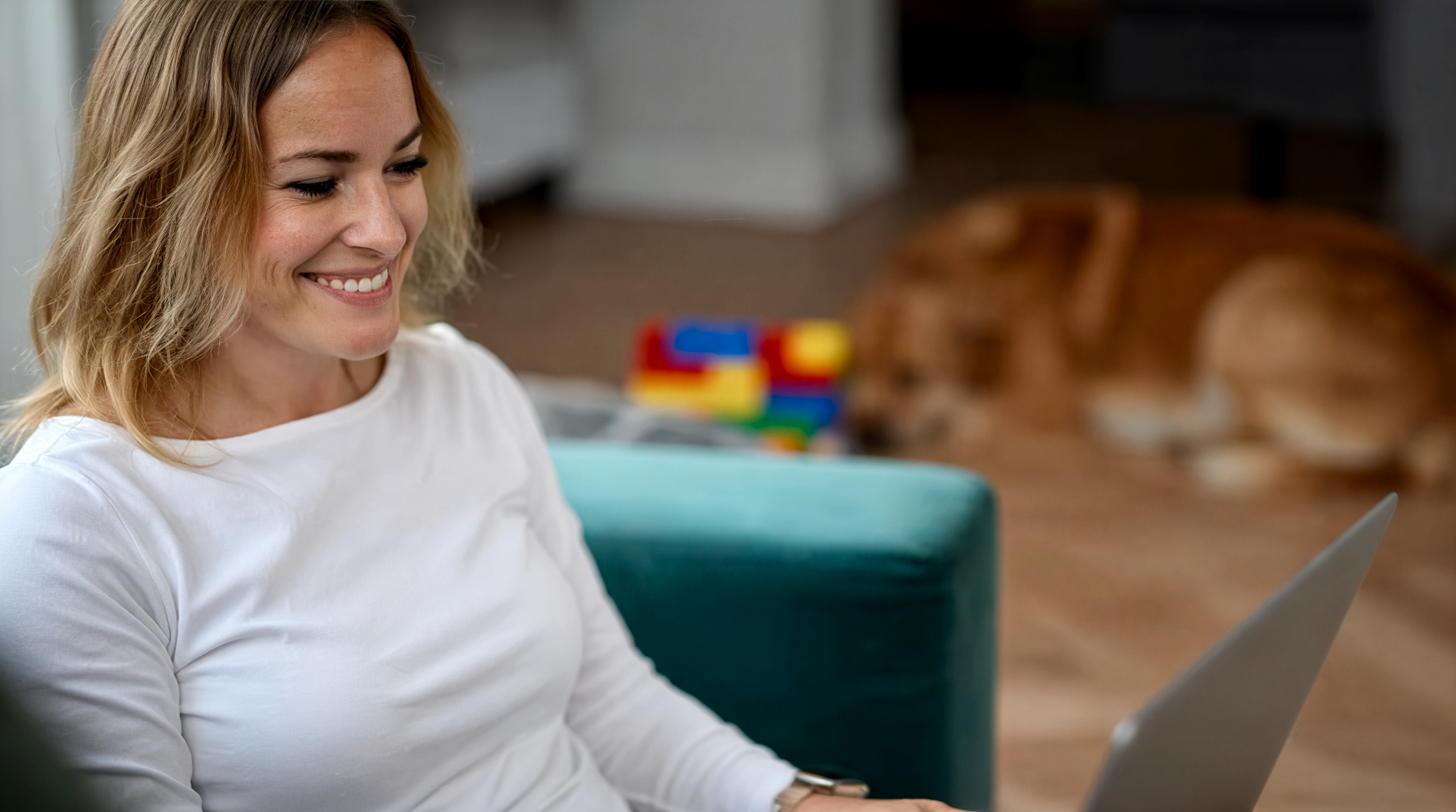 Eine lächelnde Frau in weißem Langarmshirt arbeitet konzentriert an einem Laptop in einem hellen Wohnraum. Im Hintergrund sind buntes Spielzeug und ein Hund auf dem Holzboden zu sehen. Die Szene vermittelt eine entspannte Atmosphäre. Dieses Bild wurde mit Hilfe von KI erstellt