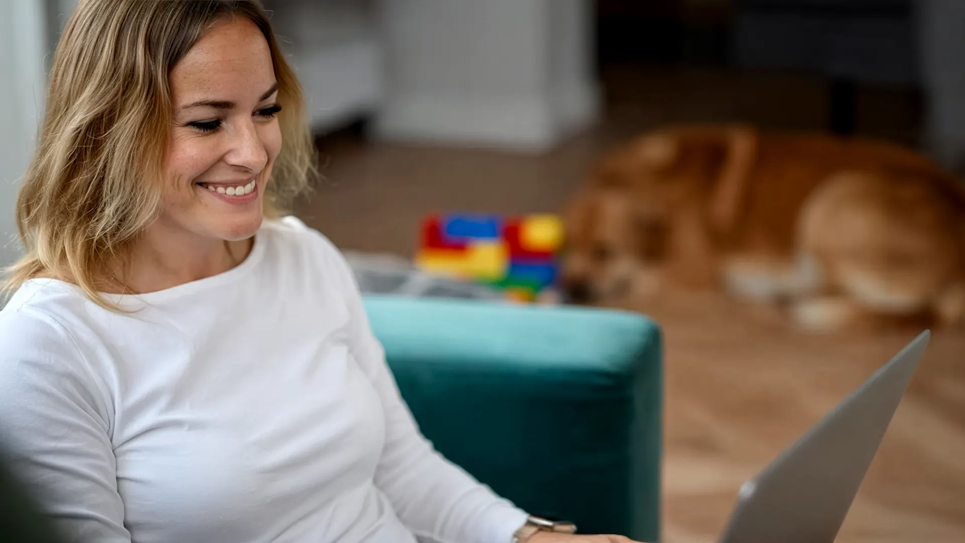 Bewerbungstipps. Dieses Bild wurde mit Hilfe von KI erstellt Eine lächelnde Frau in weißem Langarmshirt arbeitet konzentriert an einem Laptop in einem hellen Wohnraum. Im Hintergrund sind buntes Spielzeug und ein Hund auf dem Holzboden zu sehen. Die Szene vermittelt eine entspannte Atmosphäre. Dieses Bild wurde mit Hilfe von KI erstellt