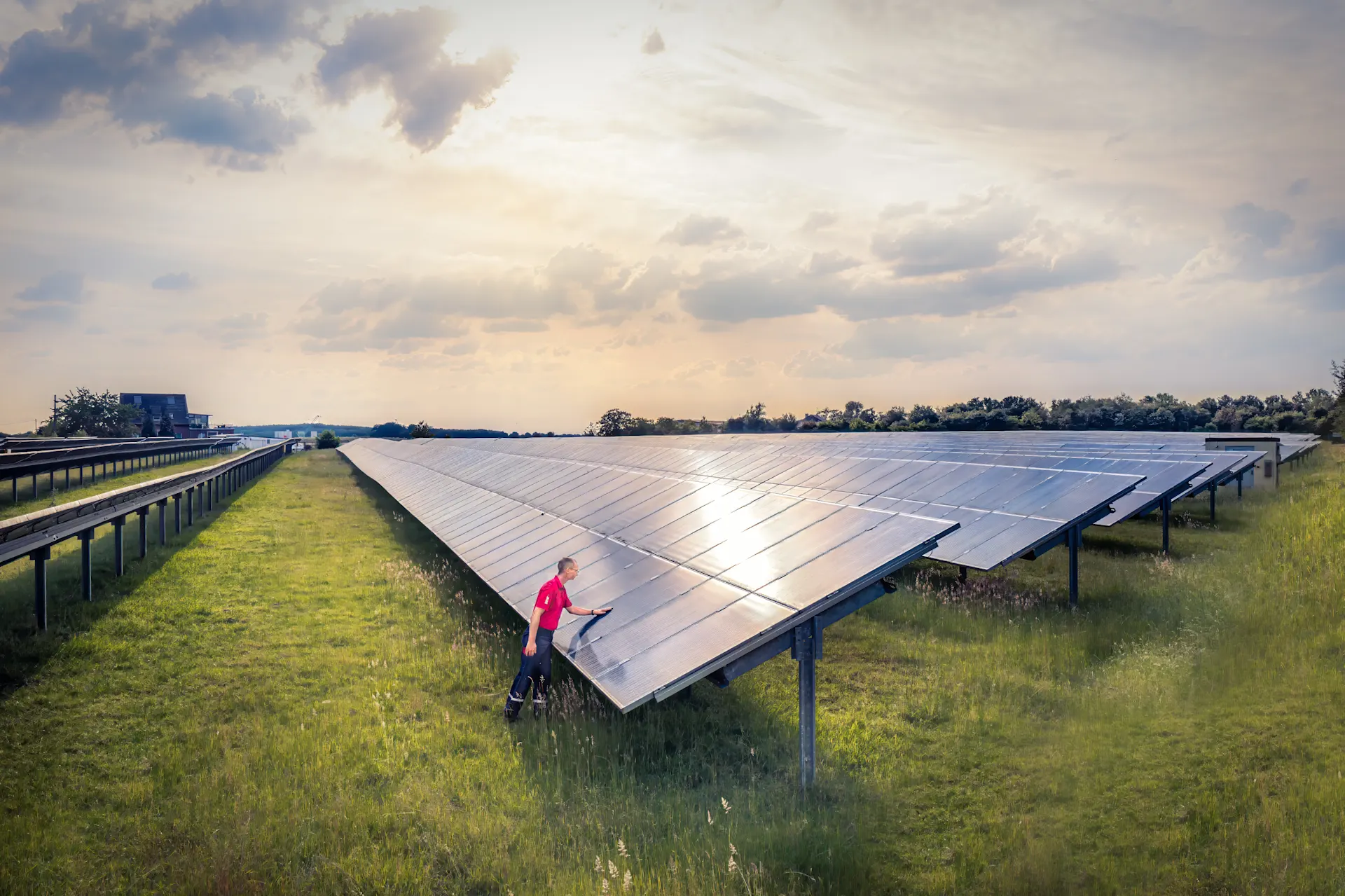 Eine Person reinigt oder inspiziert eine große Freiflächen-Photovoltaikanlage. Die Solarmodule erstrecken sich über eine Wiese bis zum Horizont, während die tief stehende Sonne durch die Wolken scheint.