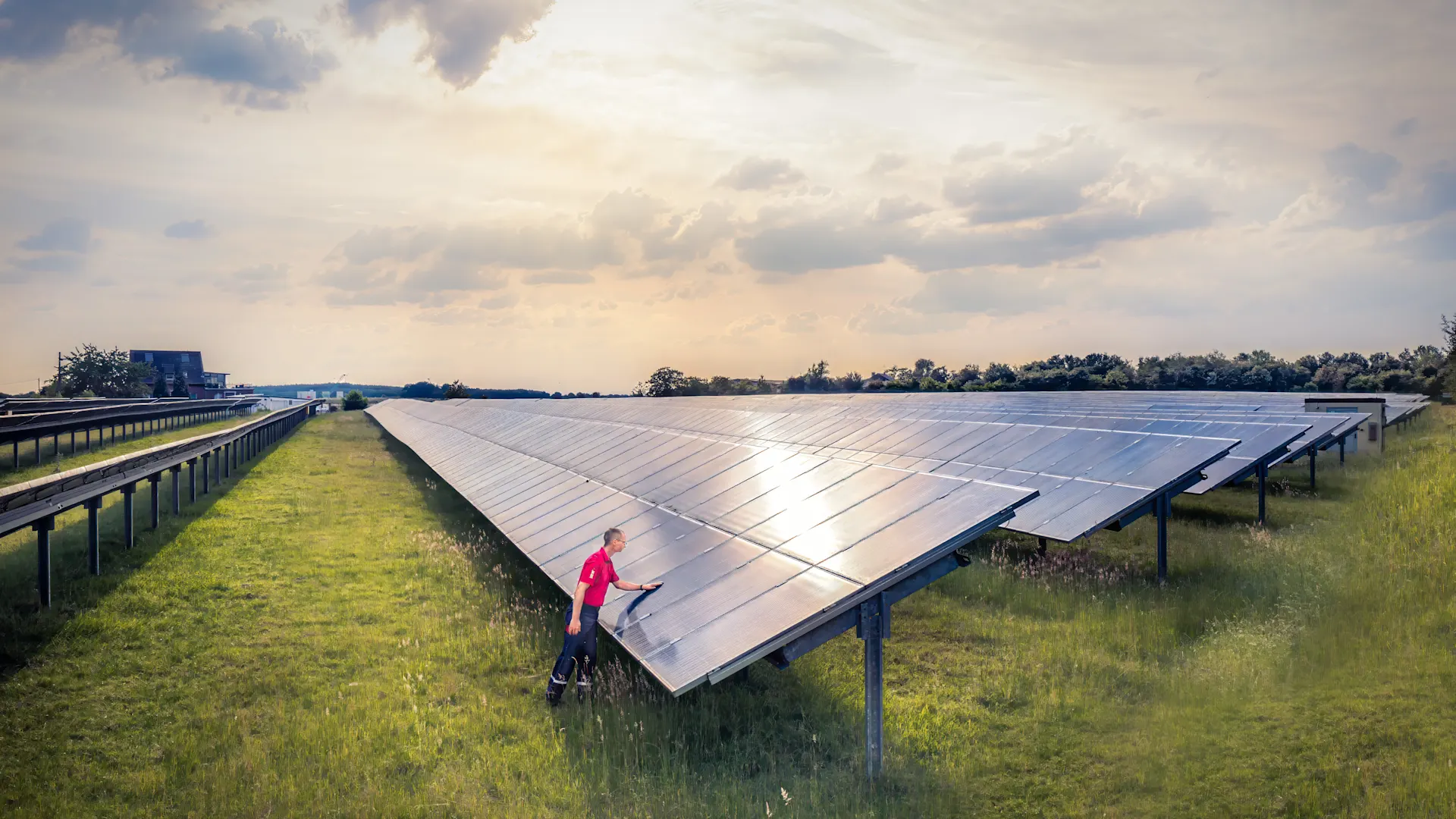 Verantwortung für Klima und Energie. Eine Person reinigt oder inspiziert eine große Freiflächen-Photovoltaikanlage. Die Solarmodule erstrecken sich über eine Wiese bis zum Horizont, während die tief stehende Sonne durch die Wolken scheint.