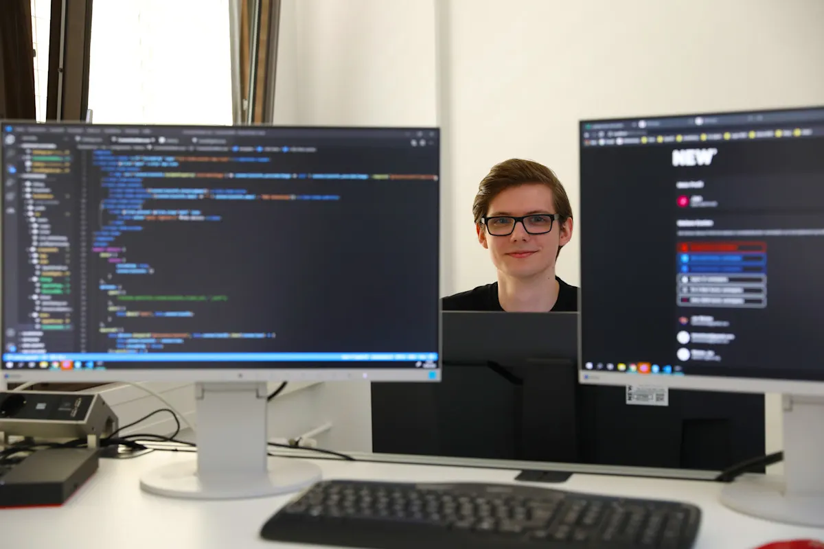 Jan Wüsten - IT-Anwendungsentwickler Moderner IT‑Arbeitsplatz im Rahmen der MATSE‑Ausbildung bei der NEW AG: Ein Schreibtisch mit zwei Monitoren zeigt auf der linken Seite Programmcode in einer Entwicklungsumgebung und auf der rechten Seite eine digitale Web‑ und Systemoberfläche im NEW‑Design. Die Arbeitsumgebung steht für praxisnahe Softwareentwicklung, moderne IT‑Ausbildung und die Verbindung von Theorie und Praxis z.B. im dualen Studiengang MATSE.