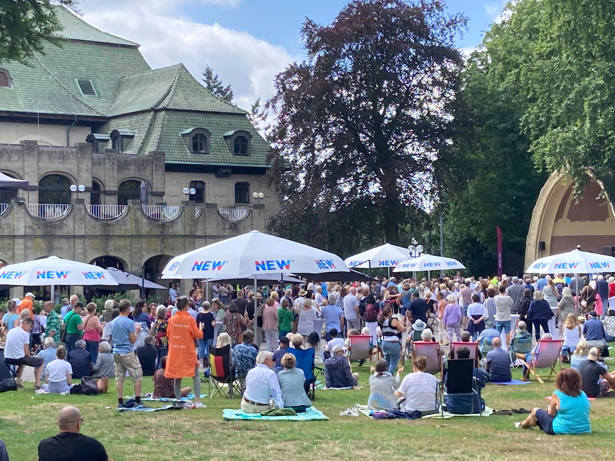 Eine große Menschenmenge versammelt sich bei einem Musikfestival im Freien vor einem Gebäude mit grünem Dach. Weiße Pavillons mit „NEW“ Beschriftung sind aufgebaut, und die Besucher:innen sitzen auf dem Rasen unter schattigen Bäumen.