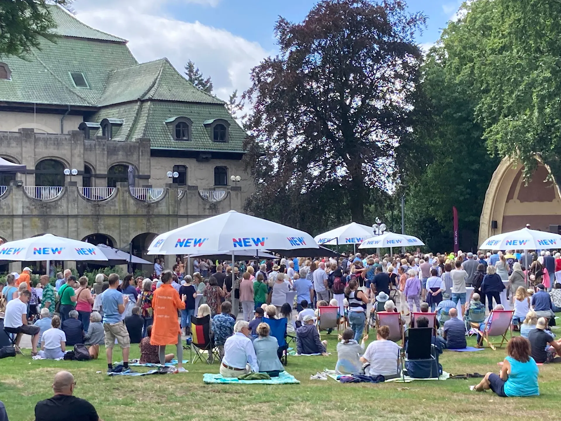 Musiksommer Eine große Menschenmenge versammelt sich bei einem Musikfestival im Freien vor einem Gebäude mit grünem Dach. Weiße Pavillons mit „NEW“ Beschriftung sind aufgebaut, und die Besucher:innen sitzen auf dem Rasen unter schattigen Bäumen.