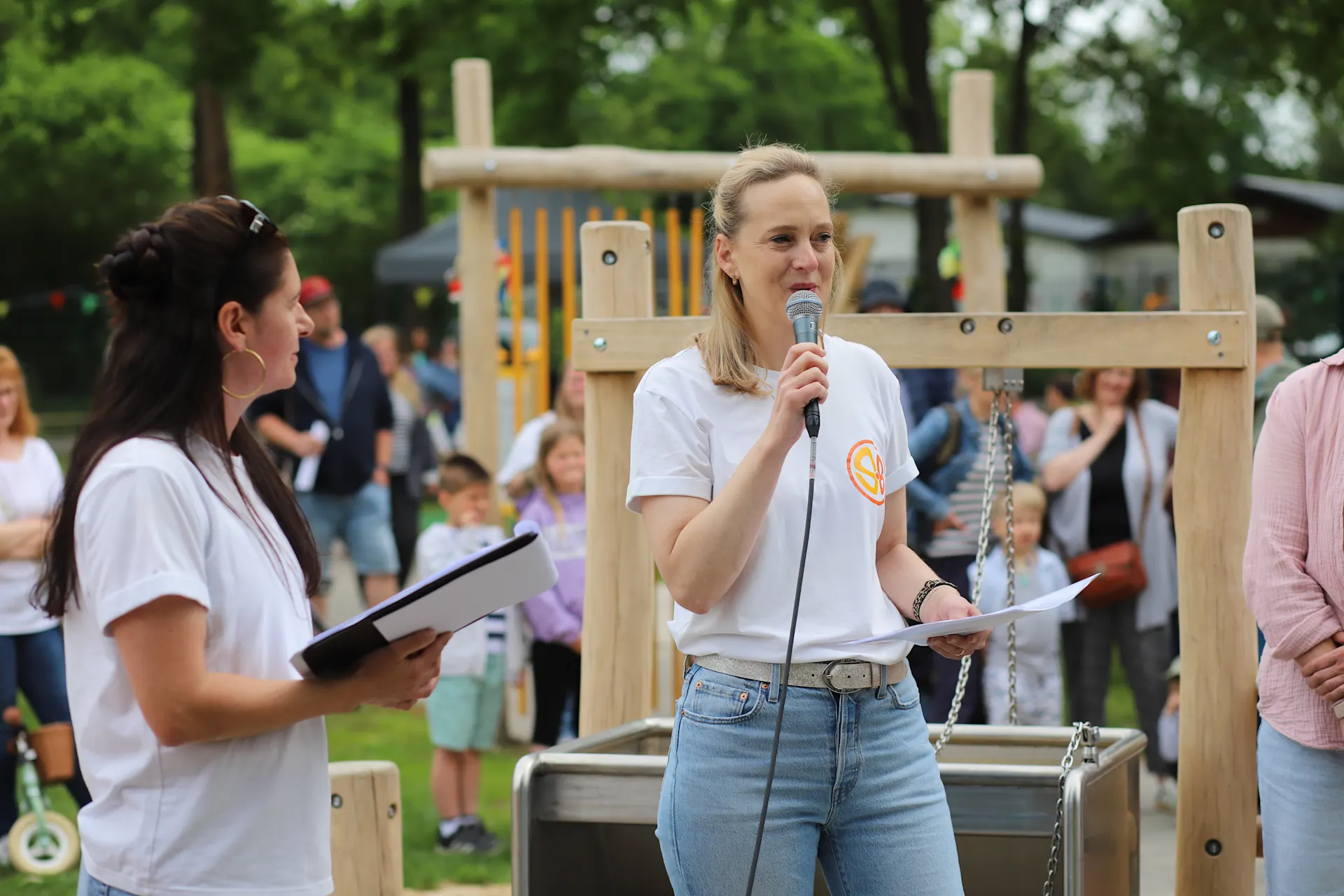 Eine Frau spricht bei der Eröffnung eines inklusiven Spielplatzes in ein Mikrofon, während eine weitere Frau mit Unterlagen daneben steht. Im Hintergrund sind Spielgeräte, Familien und Gäste der Veranstaltung zu sehen. 