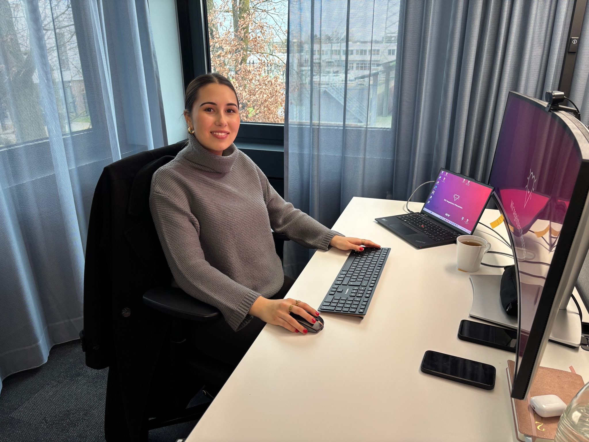Eine junge Frau sitzt lächelnd an einem modernen Büroarbeitsplatz vor einem großen Monitor und einem Laptop. Sie arbeitet in einer hellen, freundlichen Umgebung mit Blick nach draußen. Auf dem Schreibtisch liegen Smartphone, Tasse und weitere Arbeitsmaterialien