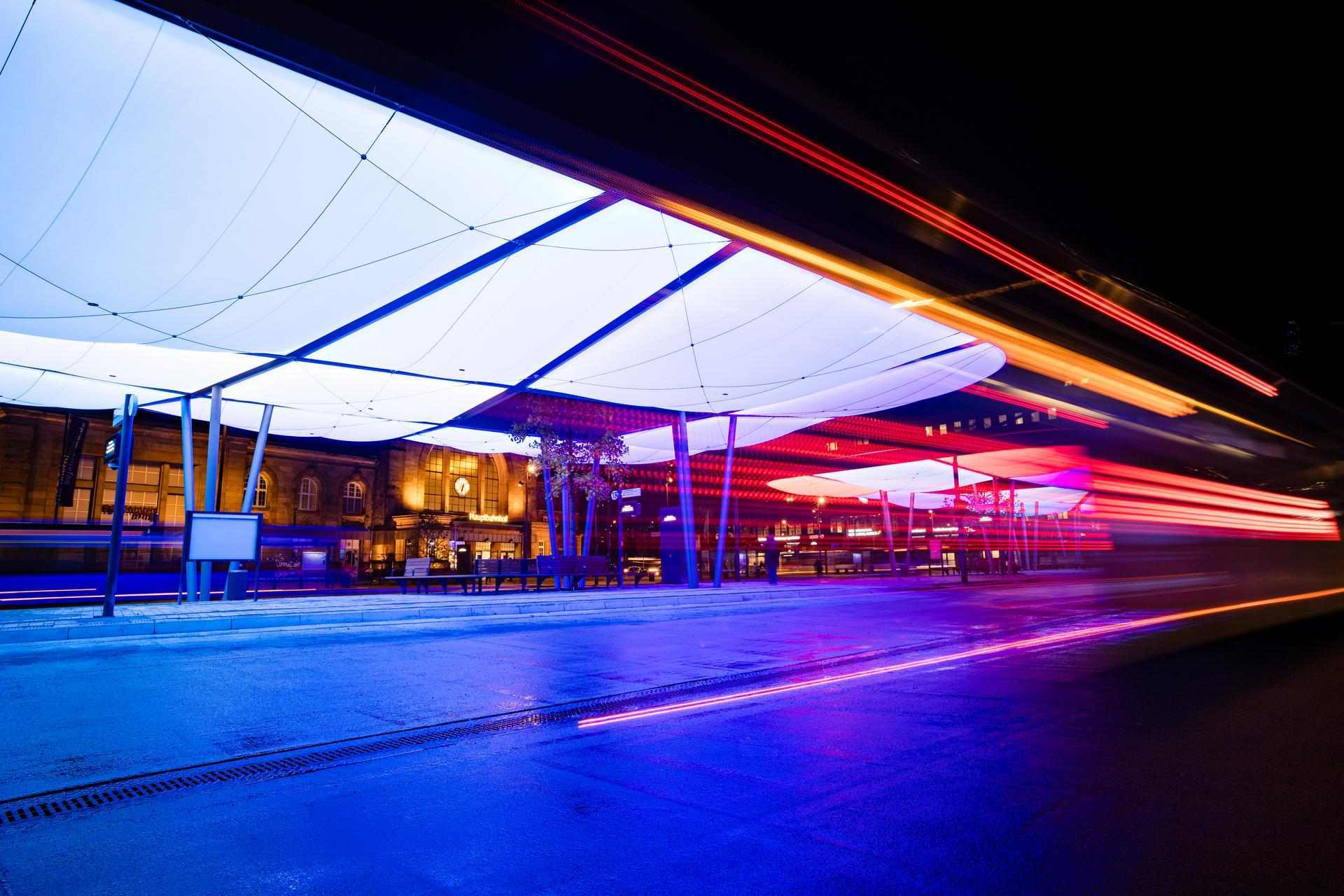 Das Bild zeigt den Zentralen Omnibusbahnhof (ZOB) in Mönchengladbach bei Nacht. Die moderne Dachkonstruktion ist farbig beleuchtet und prägt das Stadtbild. Im Hintergrund befindet sich der Hauptbahnhof. Die Szene wirkt dynamisch durch Lichtspuren vorbeifahrender Fahrzeuge.