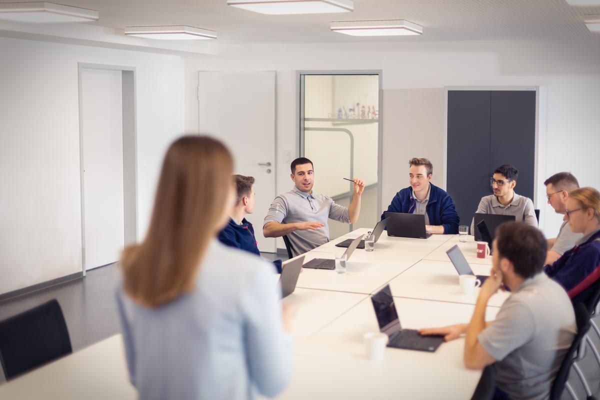 Mehrere junge Erwachsene sitzen in einem modernen Besprechungsraum um einen Konferenztisch und diskutieren angeregt, während eine Person in der Unschärfe im Vordergrund steht und zuhört.
