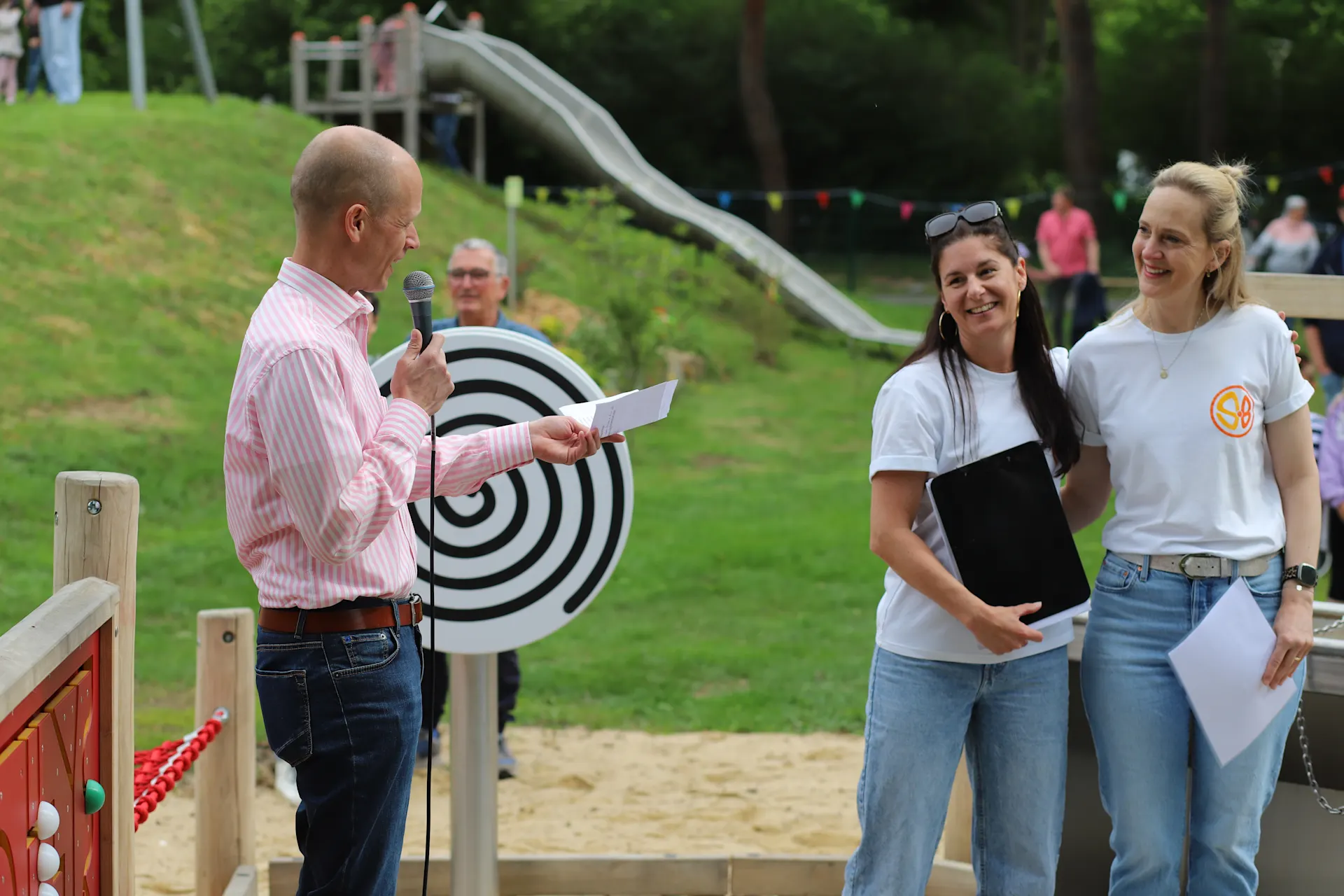 Zwei Frauen stehen lächelnd auf einem inklusiven Spielplatz neben einem Mann mit Mikrofon und Papier in der Hand, der offenbar eine Rede hält. Im Hintergrund sind Spielgeräte, eine Rutsche sowie weitere Gäste der Spielplatzeröffnung zu sehen.