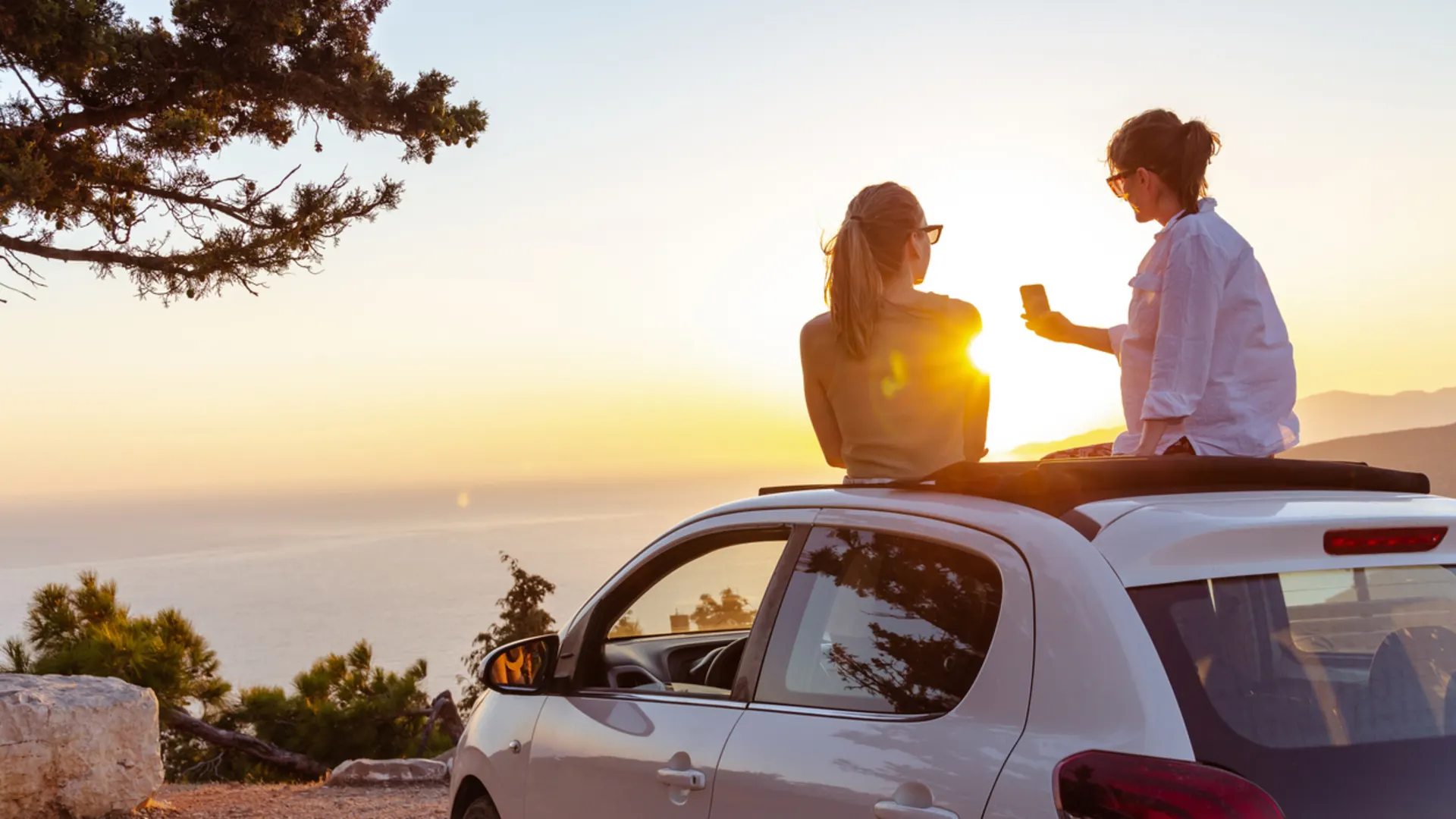Urlaub mit E-Auto: stressfrei reisen mit Ladetipps, Routen & Zielen (Foto: iStock / milko) Zwei Personen sitzen bei Sonnenuntergang auf dem Dach eines weißen Autos. Eine Person hält ein Handy in der Hand und sie genießen die Aussicht auf die Küste bei Sonnenuntergang.