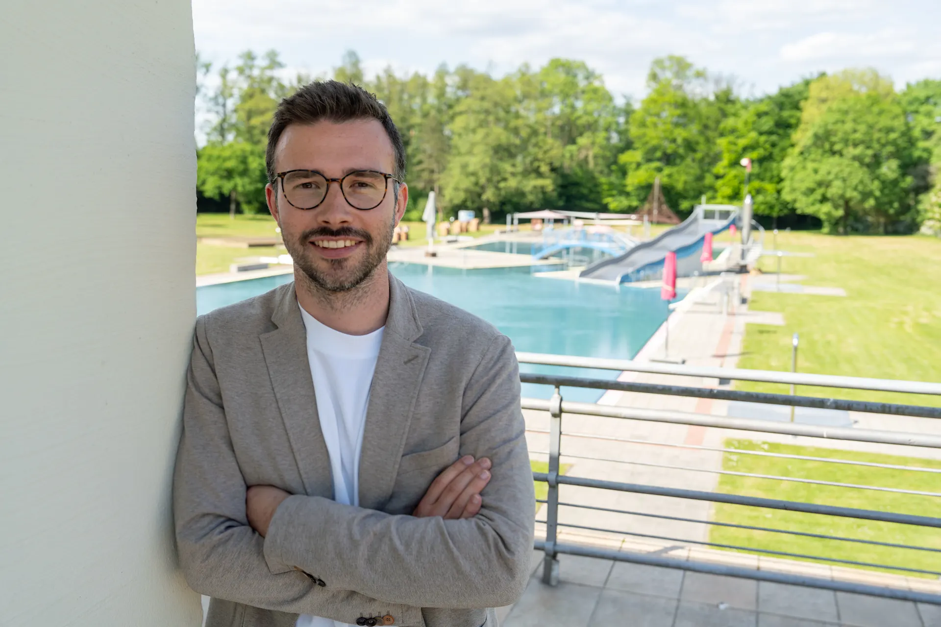 Armin Brückner Porträtaufnahme einer Person mit verschränkten Armen auf einer Terrasse. Die Person trägt ein helles Sakko über einem weißen T-Shirt und eine Brille. Im Hintergrund befindet sich ein Freibad mit mehreren Schwimmbecken, Liegewiesen und Spiel- beziehungsweise Freizeitelementen. Hinter dem Freibad ist eine Baumreihe zu sehen. Die Aufnahme entstand bei Tageslicht und zeigt die Person im Vordergrund vor der Freibadanlage.