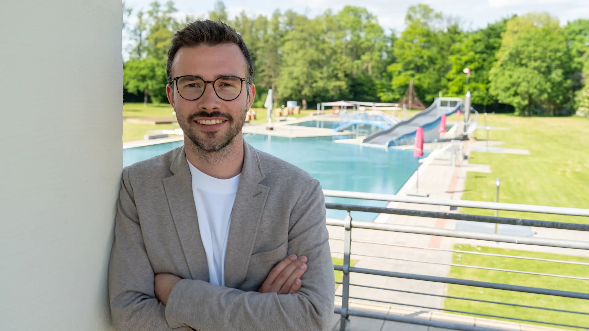 Armin Brückner Porträtaufnahme einer Person mit verschränkten Armen auf einer Terrasse. Die Person trägt ein helles Sakko über einem weißen T-Shirt und eine Brille. Im Hintergrund befindet sich ein Freibad mit mehreren Schwimmbecken, Liegewiesen und Spiel- beziehungsweise Freizeitelementen. Hinter dem Freibad ist eine Baumreihe zu sehen. Die Aufnahme entstand bei Tageslicht und zeigt die Person im Vordergrund vor der Freibadanlage.