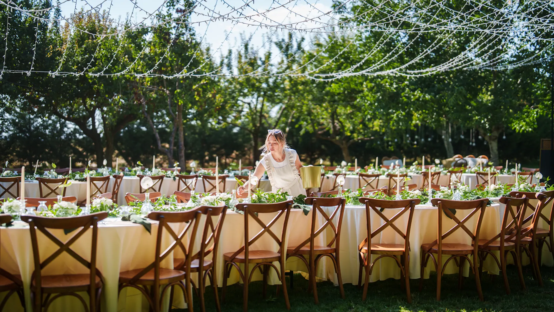 NEW Magazin Hochzeitslocations (Foto: iStock / Ugur Karakoc) Zwei große lange Tischreihen stehen in einem sonnigen Garten mit Bäumen. Eine Frau deckt ein. Die Tische sind festlich geschmückt, Lichterketten hängen darüber. Credit: Ugur Karakoc