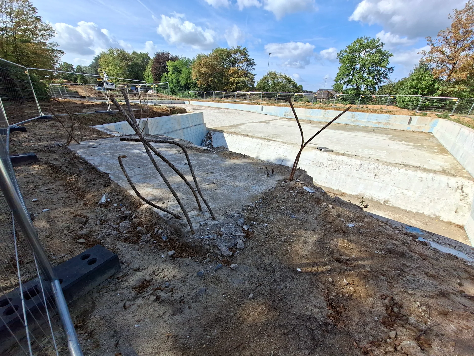 Leeres Schwimmbecken eines Freibads während der Sanierung mit Betonflächen, Baustellenabsicherung und Bäumen im Hintergrund.
