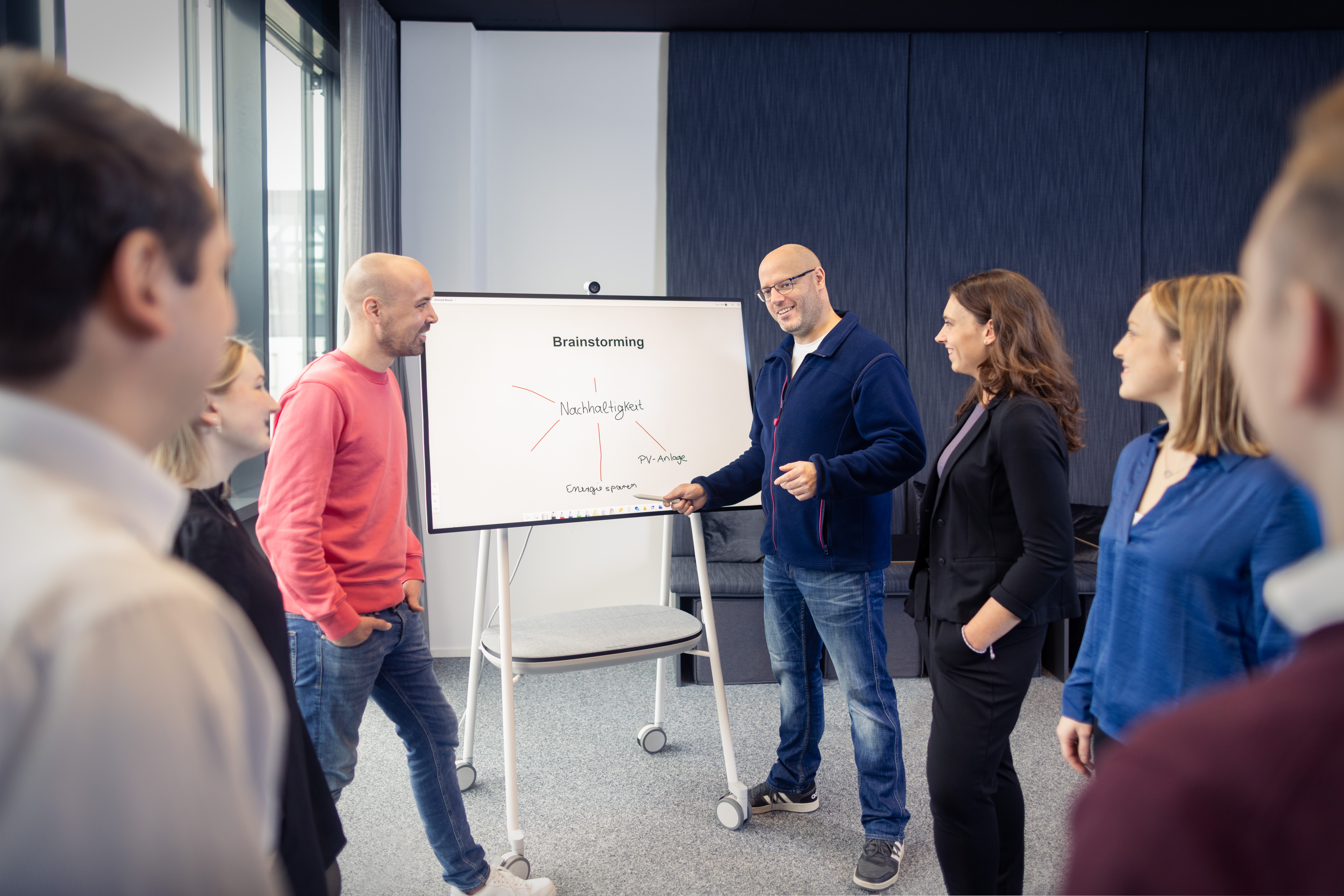 In einem hellen Büroraum mit großen Fenstern präsentiert ein Mann in dunklem Pullover vor drei Kolleg:innen an einem Whiteboard. Die Zuhörer:innen, ein Mann in rotem Pullover, eine blonde Frau und eine Frau mit braunen Haaren in dunkler Kleidung, stehen aufmerksam um das Präsentationsbrett herum.