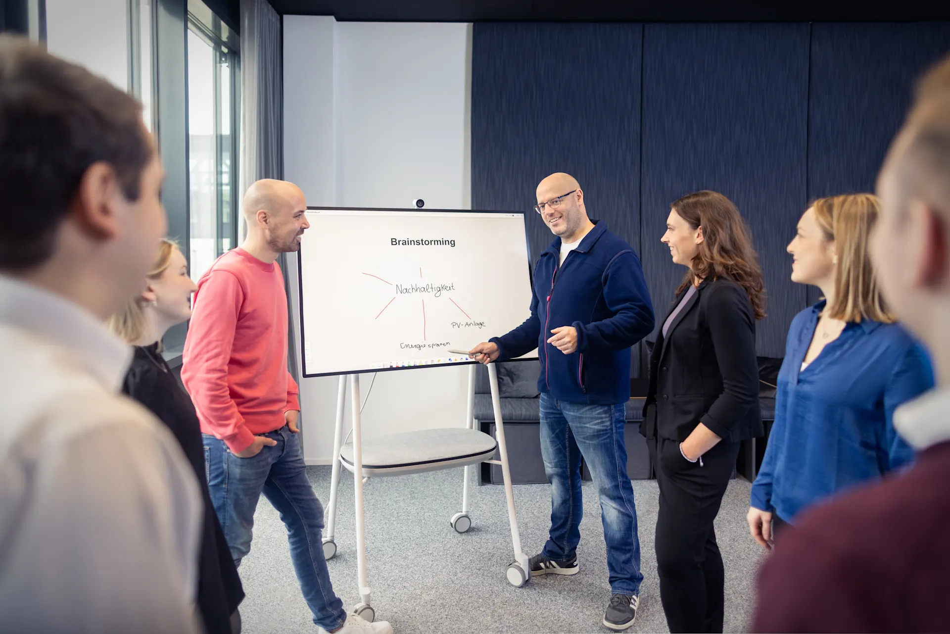 In einem hellen Büroraum mit großen Fenstern präsentiert ein Mann in dunklem Pullover vor drei Kolleg:innen an einem Whiteboard. Die Zuhörer:innen, ein Mann in rotem Pullover, eine blonde Frau und eine Frau mit braunen Haaren in dunkler Kleidung, stehen aufmerksam um das Präsentationsbrett herum.
