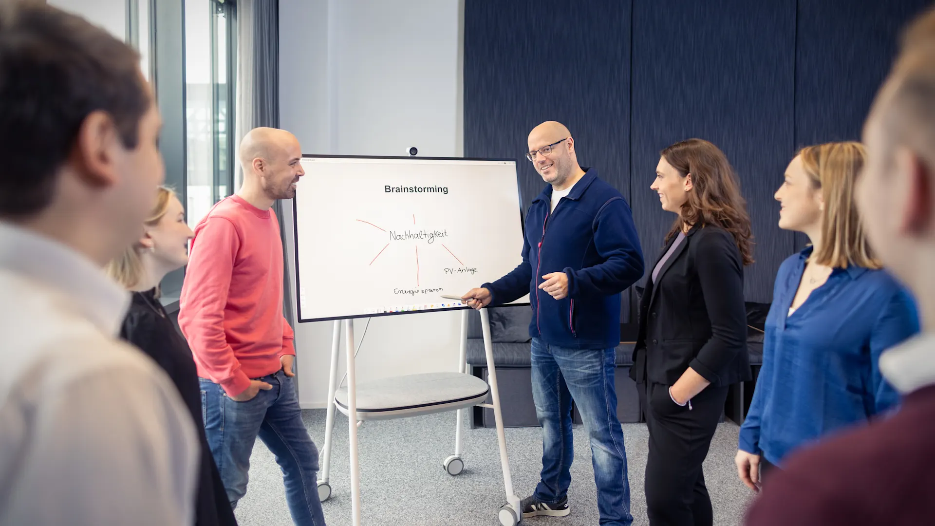 Führen mit Werten, Handeln mit Weitsicht. In einem hellen Büroraum mit großen Fenstern präsentiert ein Mann in dunklem Pullover vor drei Kolleg:innen an einem Whiteboard. Die Zuhörer:innen, ein Mann in rotem Pullover, eine blonde Frau und eine Frau mit braunen Haaren in dunkler Kleidung, stehen aufmerksam um das Präsentationsbrett herum.