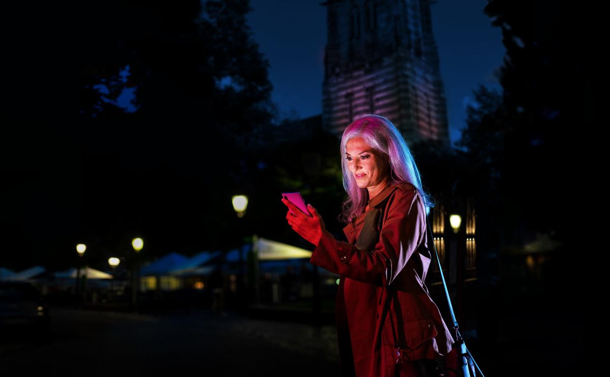 Das Bild zeigt eine junge Frau, die im Dunkeln auf einer Straße unterwegs ist. Sie trägt eine braune Jacke und hält ein Mobiltelefon in der Hand. Im Hintergrund sind Häuser, Laternen und Dekorationen zu sehen.
