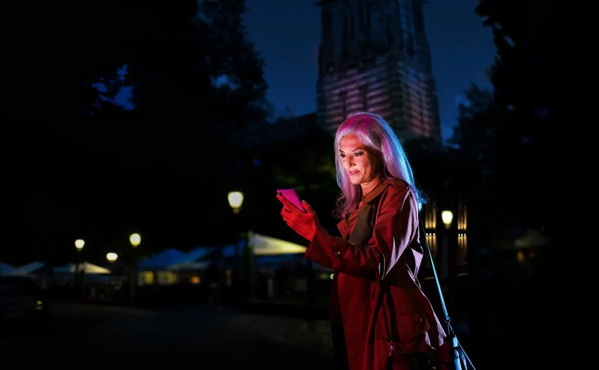 Das Bild zeigt eine junge Frau, die im Dunkeln auf einer Straße unterwegs ist. Sie trägt eine braune Jacke und hält ein Mobiltelefon in der Hand. Im Hintergrund sind Häuser, Laternen und Dekorationen zu sehen.