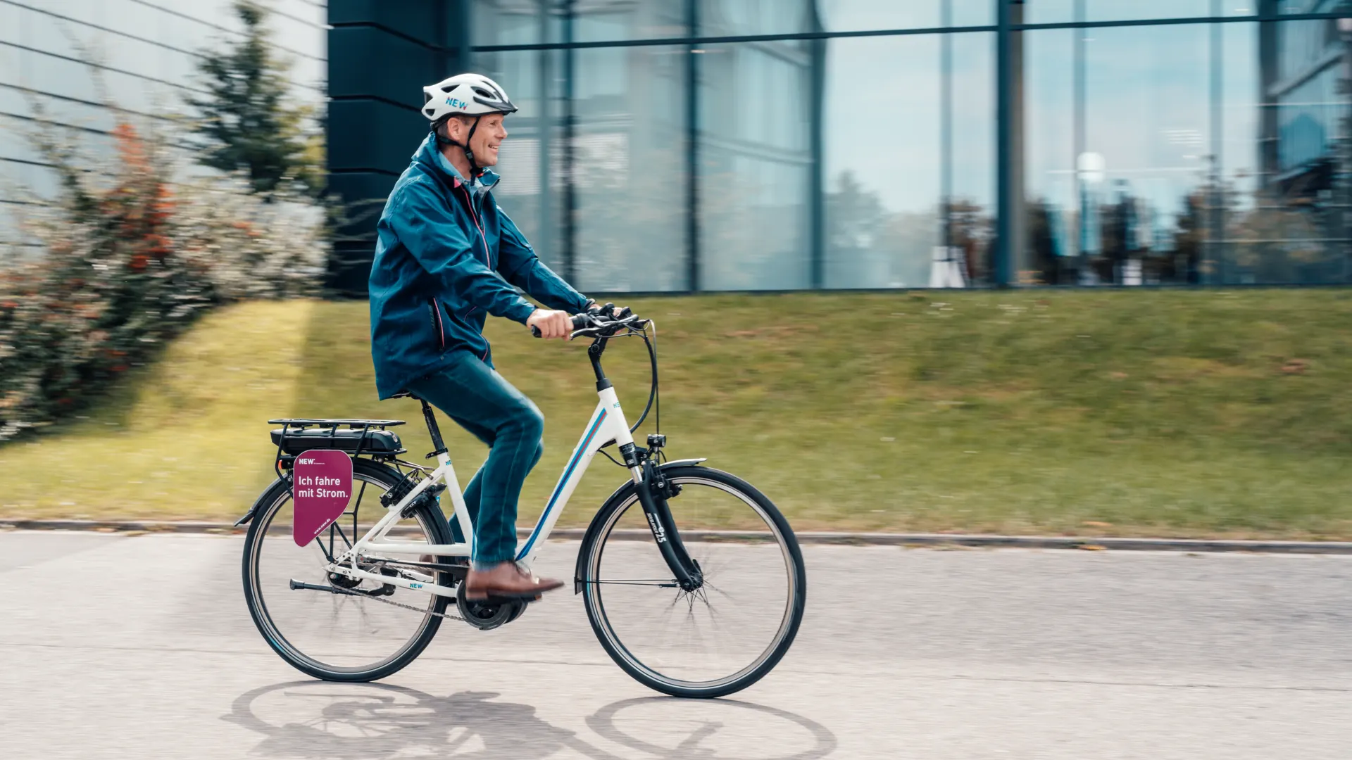 Eine Person fährt mit einem E-Bike auf einer Straße an einem modernen Gebäude mit großer Glasfassade vorbei. Die Person trägt einen weißen Fahrradhelm sowie eine dunkelblaue Jacke und Hose. Am hinteren Gepäckträger des Fahrrads befindet sich eine Fahrradtasche mit einem pinkfarbenen Motiv und der Aufschrift „Ich fahre mit Strom“. Im Hintergrund sind eine Grünfläche, Sträucher und Bäume zu sehen. Die leichte Bewegungsunschärfe im Hintergrund vermittelt den Eindruck einer Fahrt.
