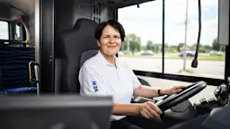 Manuela am Steuer eines Linienbusses. Dieses Bild wurde mit Hilfe von KI erstellt Eine freundlich lächelnde Busfahrerin in weißem Poloshirt sitzt am Steuer eines modernen Busses. Durch die großen Fenster neben ihr ist eine sonnige Straßenlandschaft mit grünen Bäumen zu sehen. Die Fahrerin trägt das NEW-Logo auf ihrer Arbeitskleidung. Dieses Bild wurde mit Hilfe von KI erstellt