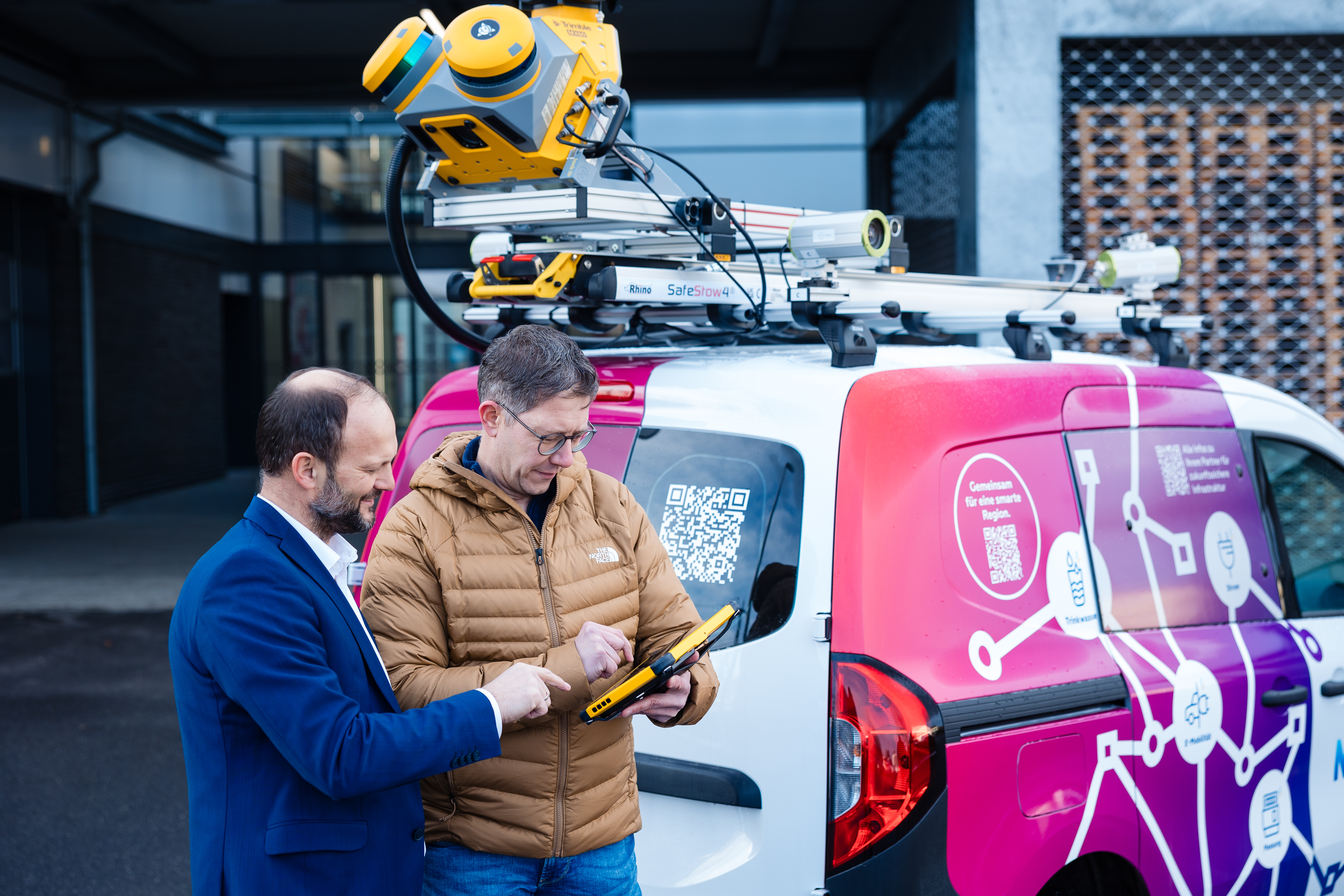 Einsatzsituation im Außenbereich: Zwei Mitarbeitende stehen neben einem Servicefahrzeug mit auffälligem NEW‑Branding und technischer Ausstattung auf dem Dach. Dort ist ein spezialisiertes Mess‑ oder Erfassungssystem montiert. Eine Person hält ein mobiles Endgerät, während die andere unterstützend auf die Anzeige zeigt. Die Szene vermittelt moderne, digitale Arbeitsprozesse, mobile Datenerfassung und den Einsatz innovativer Technik im praktischen Einsatzfeld.