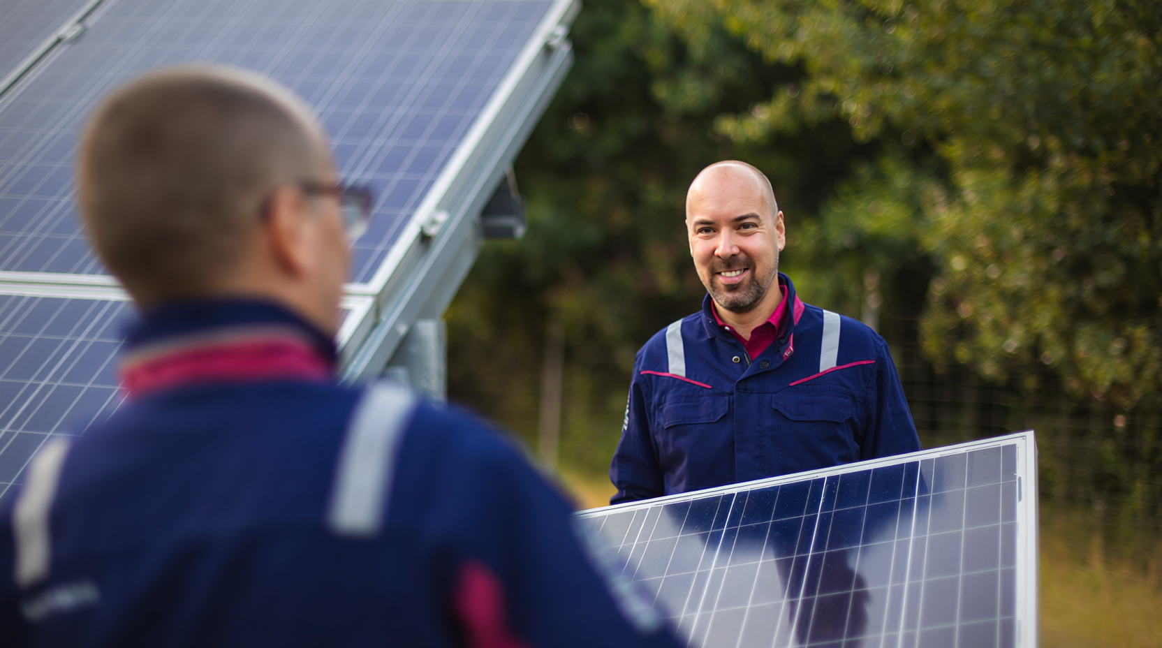 Zwei Techniker in blauer Arbeitskleidung mit reflektierenden Streifen arbeiten gemeinsam an der Installation von Solarmodulen im Freien. Im Hintergrund sind weitere Solarpaneele und grüne Bäume zu sehen. Dieses Bild wurde mit Hilfe von KI erstellt