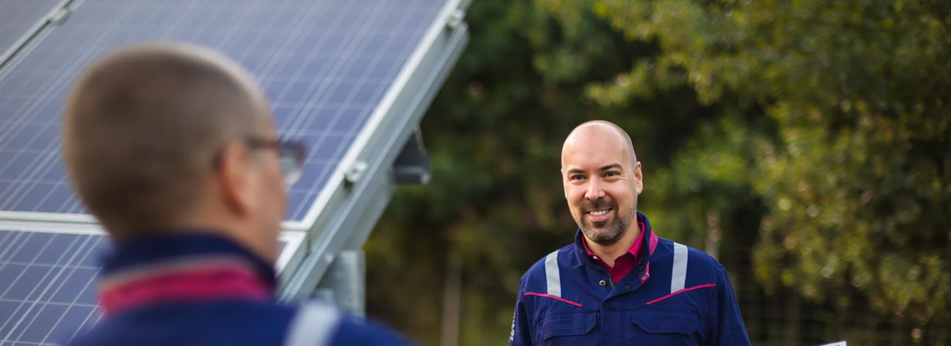 Berufseinssteiger - Zwei Mitarbeiter in einem Solarpark. Dieses Bild wurde mit Hilfe von KI erstellt Zwei Techniker in blauer Arbeitskleidung mit reflektierenden Streifen arbeiten gemeinsam an der Installation von Solarmodulen im Freien. Im Hintergrund sind weitere Solarpaneele und grüne Bäume zu sehen. Dieses Bild wurde mit Hilfe von KI erstellt