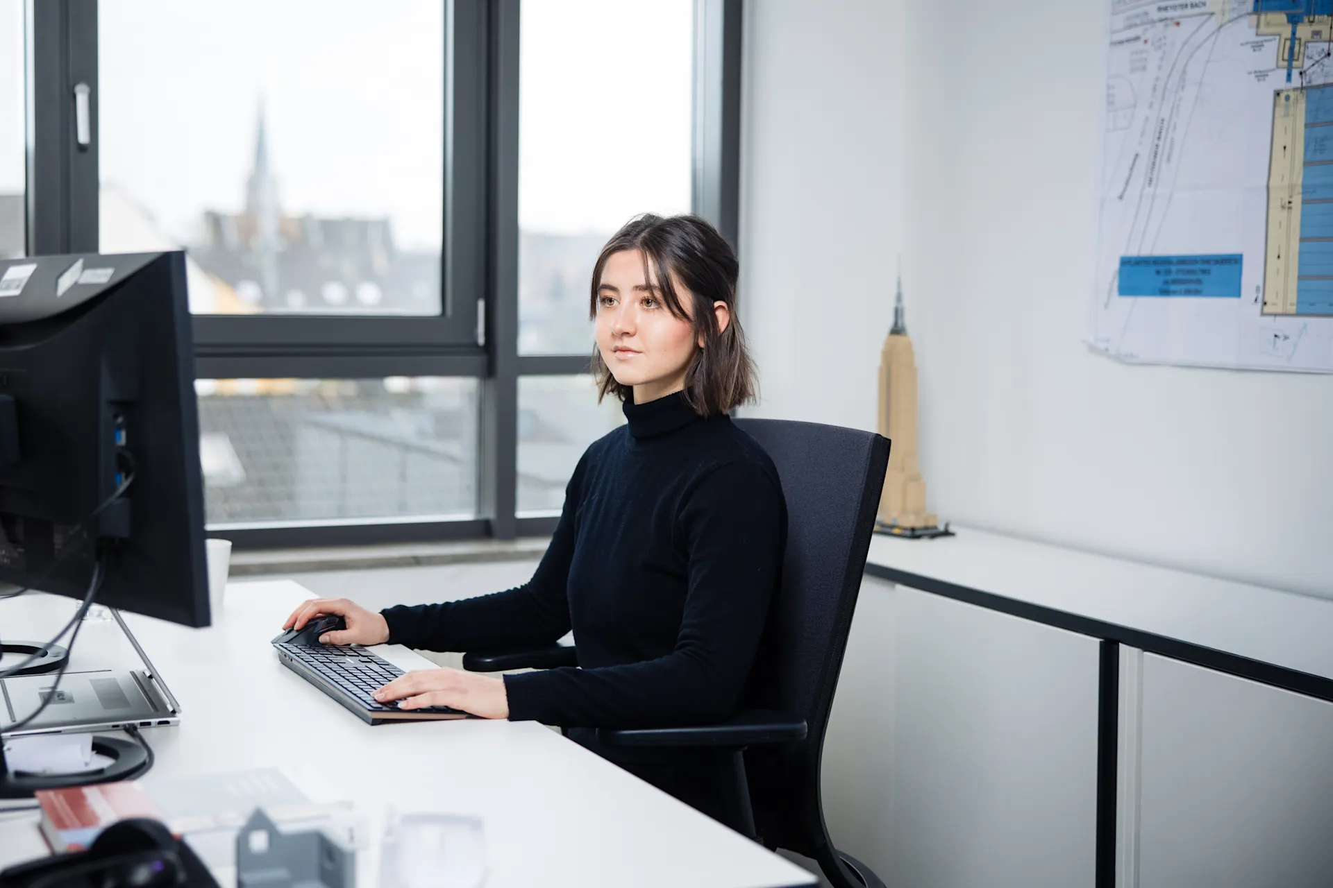 Von Buenos Aires zur Ausbildung bei der NEW Eine junge Frau sitzt in einem Büro an einem Schreibtisch. Sie schaut konzentriert auf einen Computerbildschirm. Im Hintergrund hängen Baupläne an der Wand.