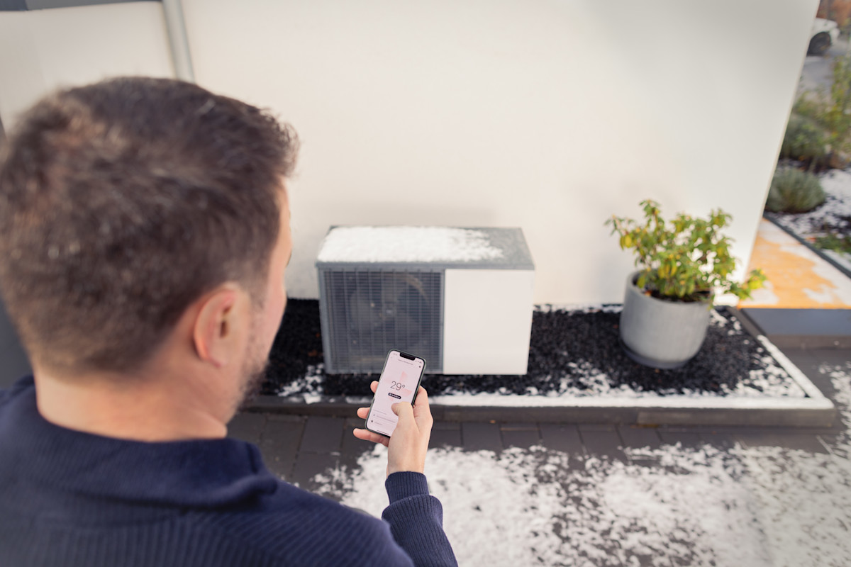 Ein Mann in dunklem Pullover bedient eine Wärmepumpe über eine Smartphone-App. Auf dem Handy-Display ist eine Temperaturanzeige sichtbar. Die moderne kleine Wärmepumpe steht auf einem Kiesbett neben einem grauen Blumentopf mit grüner Pflanze.