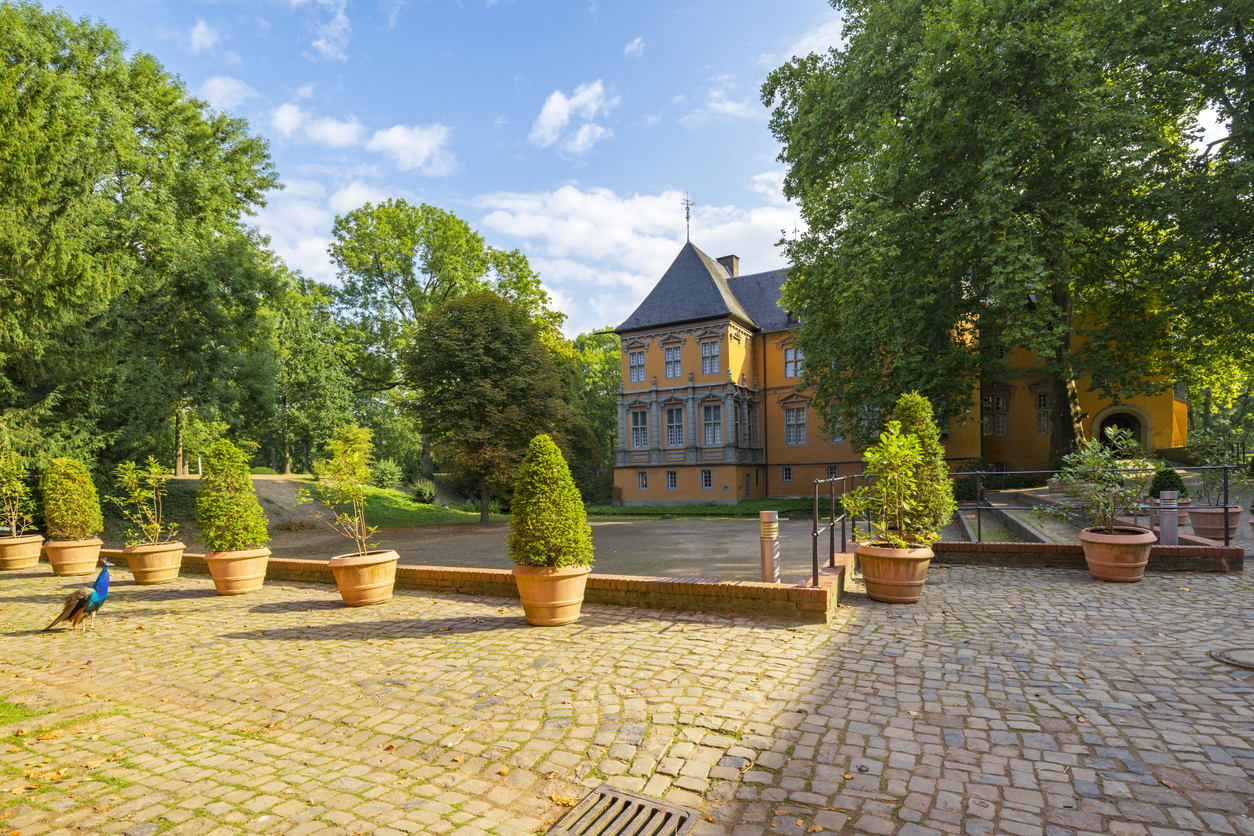 Gepflasterter Innenhof eines historischen orangefarbenen Gebäudes, umgeben von Laubbäumen. Im Vordergrund ist ein Pfau zu erkennen. Der Himmel ist blau mit einzelnen Wolken.  