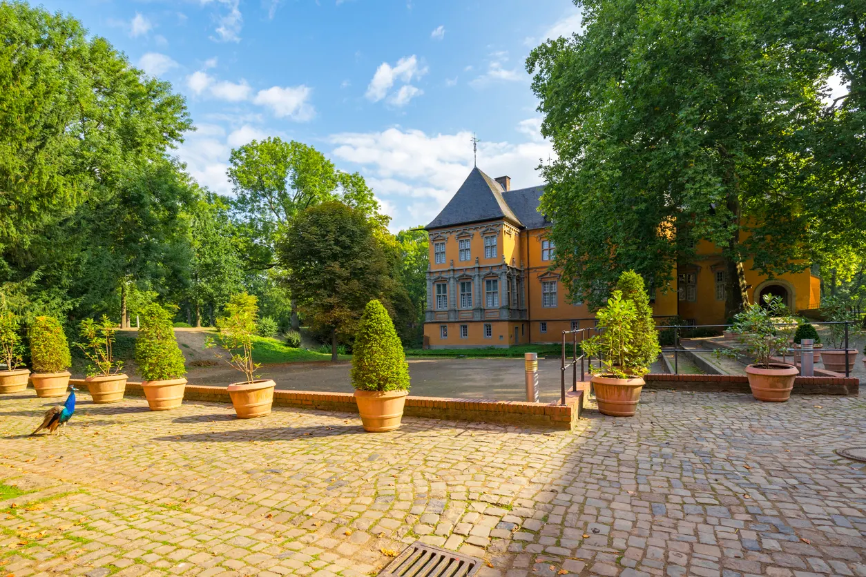 Kunst und Kultur in Mönchengladbach | iStock/ Manninx Gepflasterter Innenhof eines historischen orangefarbenen Gebäudes, umgeben von Laubbäumen. Im Vordergrund ist ein Pfau zu erkennen. Der Himmel ist blau mit einzelnen Wolken.