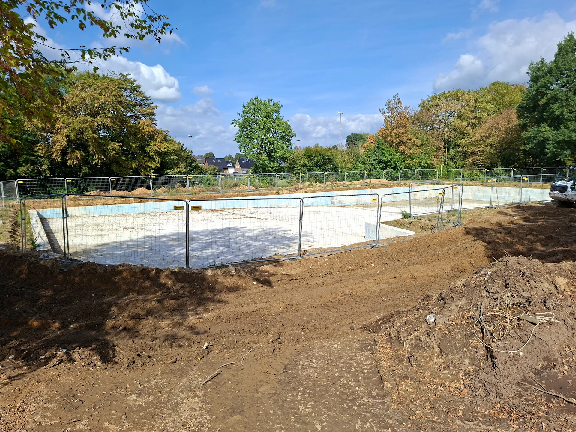Leeres Schwimmbecken eines Freibads während der Sanierung mit Betonflächen, Baustellenabsicherung und Bäumen im Hintergrund.