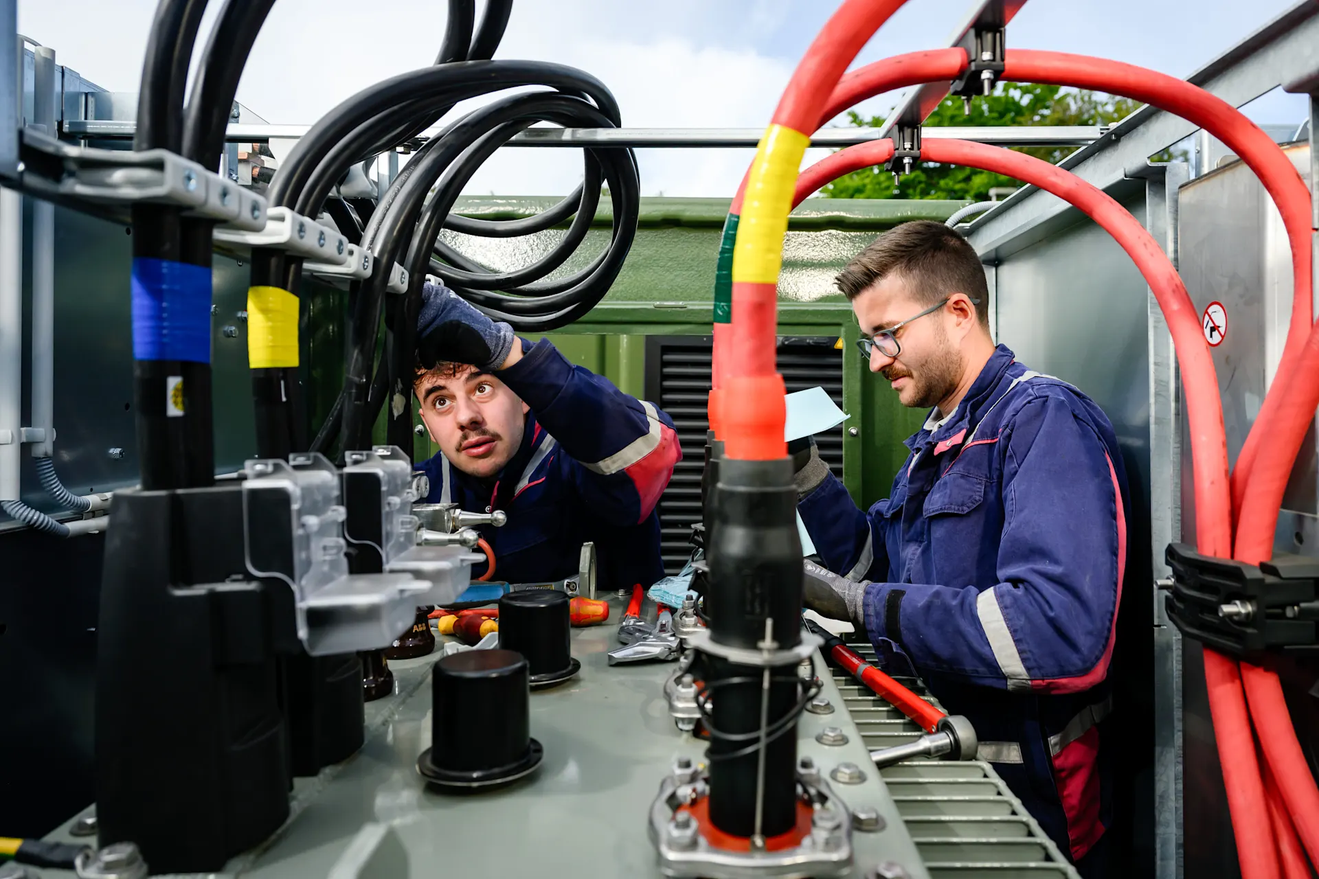 Monteur:in: handwerklicher Beruf mit Zukunft Zwei Fachkräfte arbeiten in einer Trafostation an der Installation und Wartung von Energiekabeln und technischen Komponenten. Sichtbar sind mehrere Stromkabel, Anschlüsse und Schaltanlagen, die für eine sichere und zuverlässige Energieversorgung sorgen. Die Aufnahme vermittelt moderne Netztechnik, Teamarbeit und die technische Expertise der NEW für eine zukunftsfähige Infrastruktur.