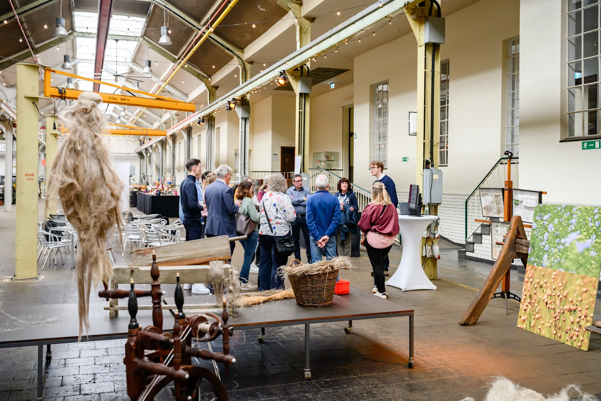 Kick-off NEW Core4Energy 14.04.2026 0176 Menschen stehen in einer lichtdurchfluteten Industriehalle zusammen und unterhalten sich. Im Vordergrund sind ein Spinnrad und Naturfasern auf einem Tisch zu sehen, im Hintergrund Stehtische und ein Gemälde mit Blumenmotiv.