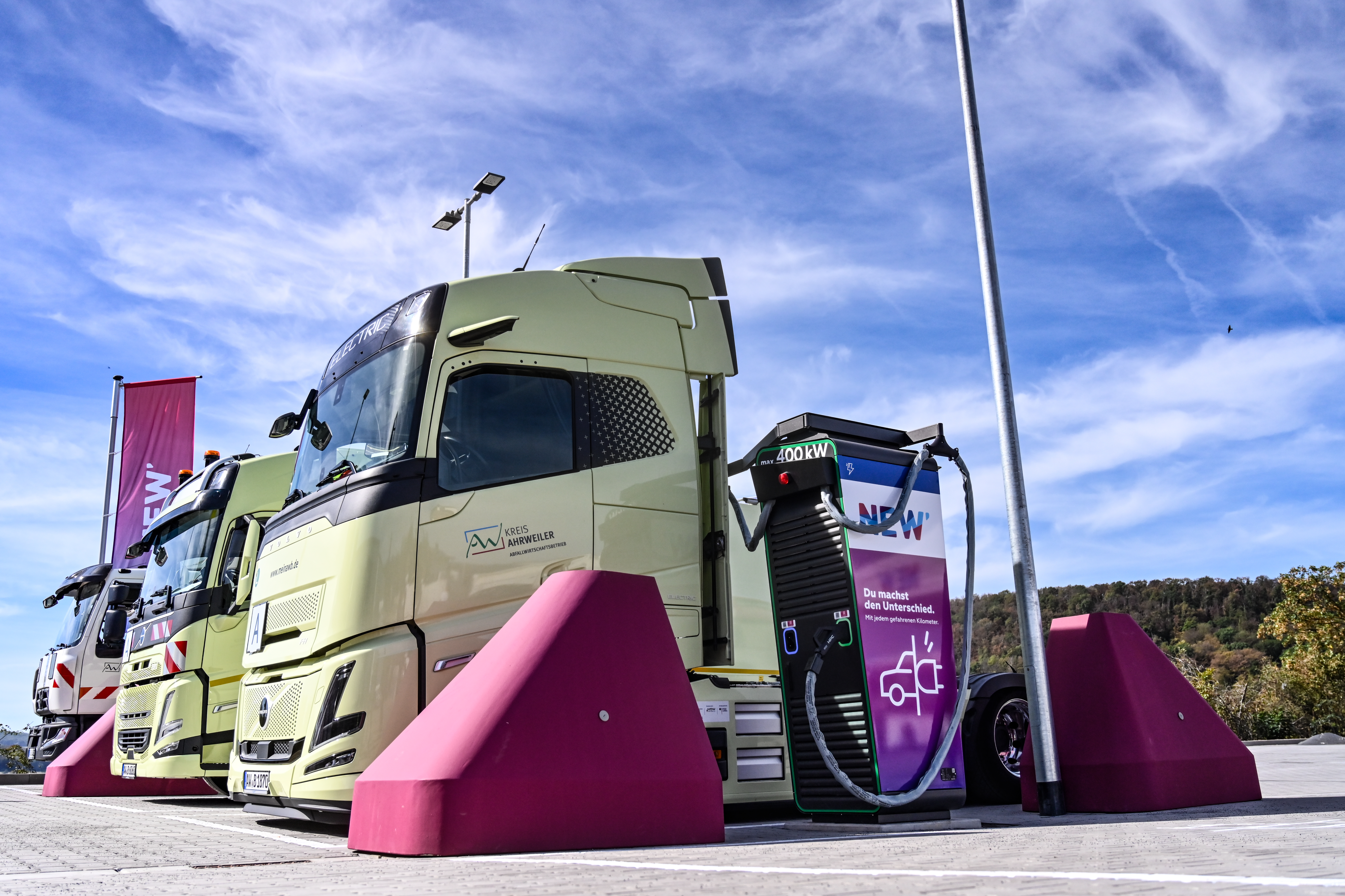 Ein hellgrüner Elektro-LKW steht an einer Schnellladestation auf einem Ladepark. Das Fahrzeug ist über ein dickes Ladekabel mit einer schwarz-violetten Ladesäule verbunden, die mit dem NEW-Logo gekennzeichnet ist. Vor der Ladesäule und neben dem Fahrzeug befinden sich auffällige magentafarbene Rammschutzblöcke. Im Hintergrund sind weitere Fahrzeuge, Fahnen und eine bewaldete Landschaft zu sehen. Über der Szene erstreckt sich ein blauer Himmel mit weißen Wolken.
