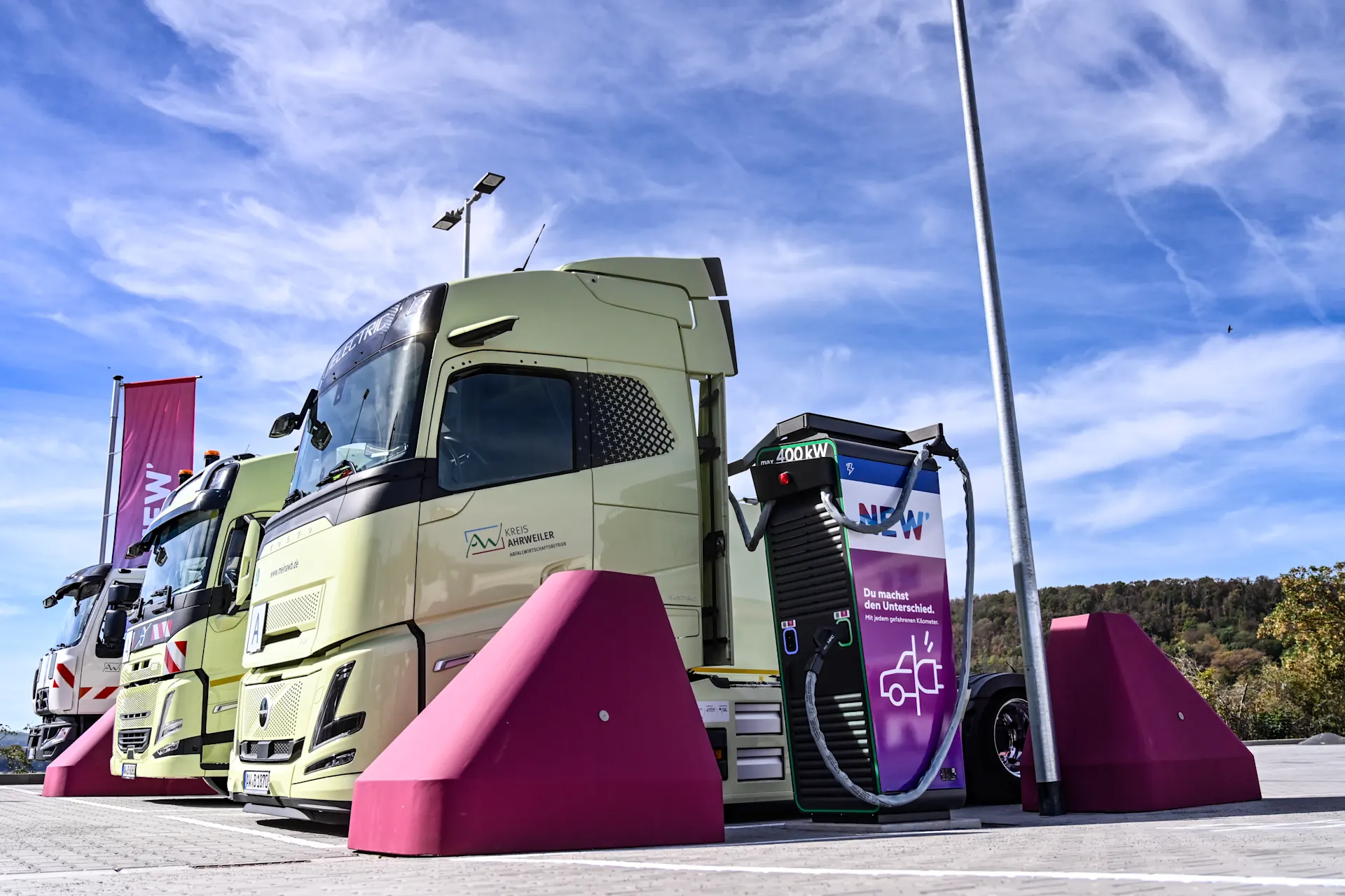 NEW Energie E-Ladepark Niederzissen Ein hellgrüner Elektro-LKW steht an einer Schnellladestation auf einem Ladepark. Das Fahrzeug ist über ein dickes Ladekabel mit einer schwarz-violetten Ladesäule verbunden, die mit dem NEW-Logo gekennzeichnet ist. Vor der Ladesäule und neben dem Fahrzeug befinden sich auffällige magentafarbene Rammschutzblöcke. Im Hintergrund sind weitere Fahrzeuge, Fahnen und eine bewaldete Landschaft zu sehen. Über der Szene erstreckt sich ein blauer Himmel mit weißen Wolken.