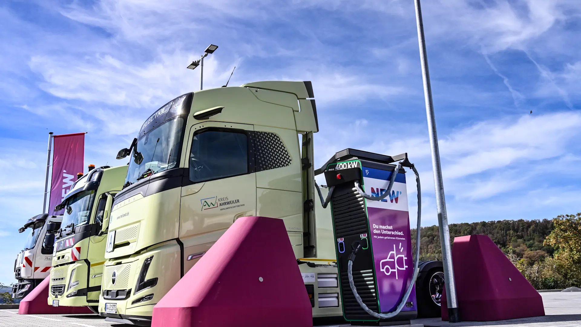 NEW Energie E-Ladepark Niederzissen Ein hellgrüner Elektro-LKW steht an einer Schnellladestation auf einem Ladepark. Das Fahrzeug ist über ein dickes Ladekabel mit einer schwarz-violetten Ladesäule verbunden, die mit dem NEW-Logo gekennzeichnet ist. Vor der Ladesäule und neben dem Fahrzeug befinden sich auffällige magentafarbene Rammschutzblöcke. Im Hintergrund sind weitere Fahrzeuge, Fahnen und eine bewaldete Landschaft zu sehen. Über der Szene erstreckt sich ein blauer Himmel mit weißen Wolken.