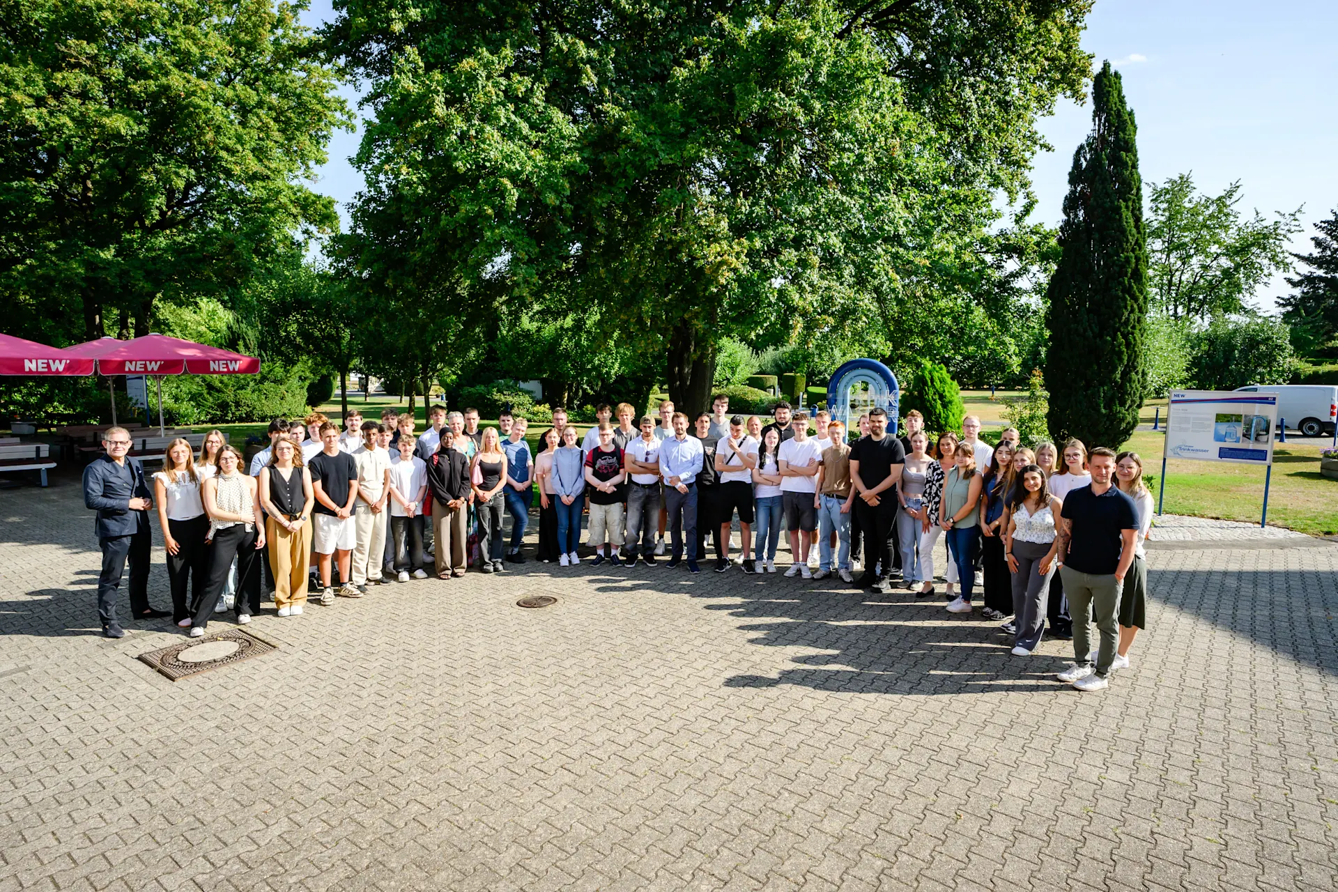 Gruppenfoto einer großen Anzahl neuer Auszubildender sowie beteiligter Mitarbeitender der NEW auf einem befestigten Platz im Außenbereich. Die Teilnehmenden stehen in mehreren Reihen vor einer grünen Park- und Gartenkulisse mit großen Bäumen. Links ist ein roter Sonnenschirm mit NEW-Branding zu sehen. Das Foto zeigt das gemeinsame Zusammenkommen im Rahmen einer Willkommens- oder Einführungsveranstaltung für den Ausbildungsstart bei der NEW. Die Aufnahme vermittelt den Gemeinschaftsgedanken, den persönlichen Austausch und den Start in die berufliche Zukunft innerhalb der NEW-Gruppe.