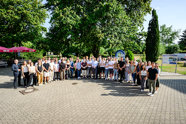 NEW Azubi Willkommen-tung 03.08.2026 Gruppenfoto einer großen Anzahl neuer Auszubildender sowie beteiligter Mitarbeitender der NEW auf einem befestigten Platz im Außenbereich. Die Teilnehmenden stehen in mehreren Reihen vor einer grünen Park- und Gartenkulisse mit großen Bäumen. Links ist ein roter Sonnenschirm mit NEW-Branding zu sehen. Das Foto zeigt das gemeinsame Zusammenkommen im Rahmen einer Willkommens- oder Einführungsveranstaltung für den Ausbildungsstart bei der NEW. Die Aufnahme vermittelt den Gemeinschaftsgedanken, den persönlichen Austausch und den Start in die berufliche Zukunft innerhalb der NEW-Gruppe.