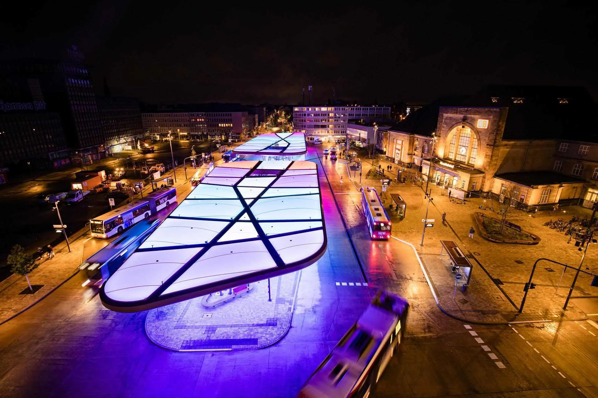 Busbahnhof Europaplatz von oben Das Bild zeigt den Zentralen Omnibusbahnhof (ZOB) in Mönchengladbach bei Nacht. Die moderne Dachkonstruktion ist farbig beleuchtet und prägt das Stadtbild. Rechts im Bild befindet sich der Hauptbahnhof, links das markante Gebäude „Haus Westland“. Die Szene wirkt dynamisch durch Lichtspuren vorbeifahrender Fahrzeuge.