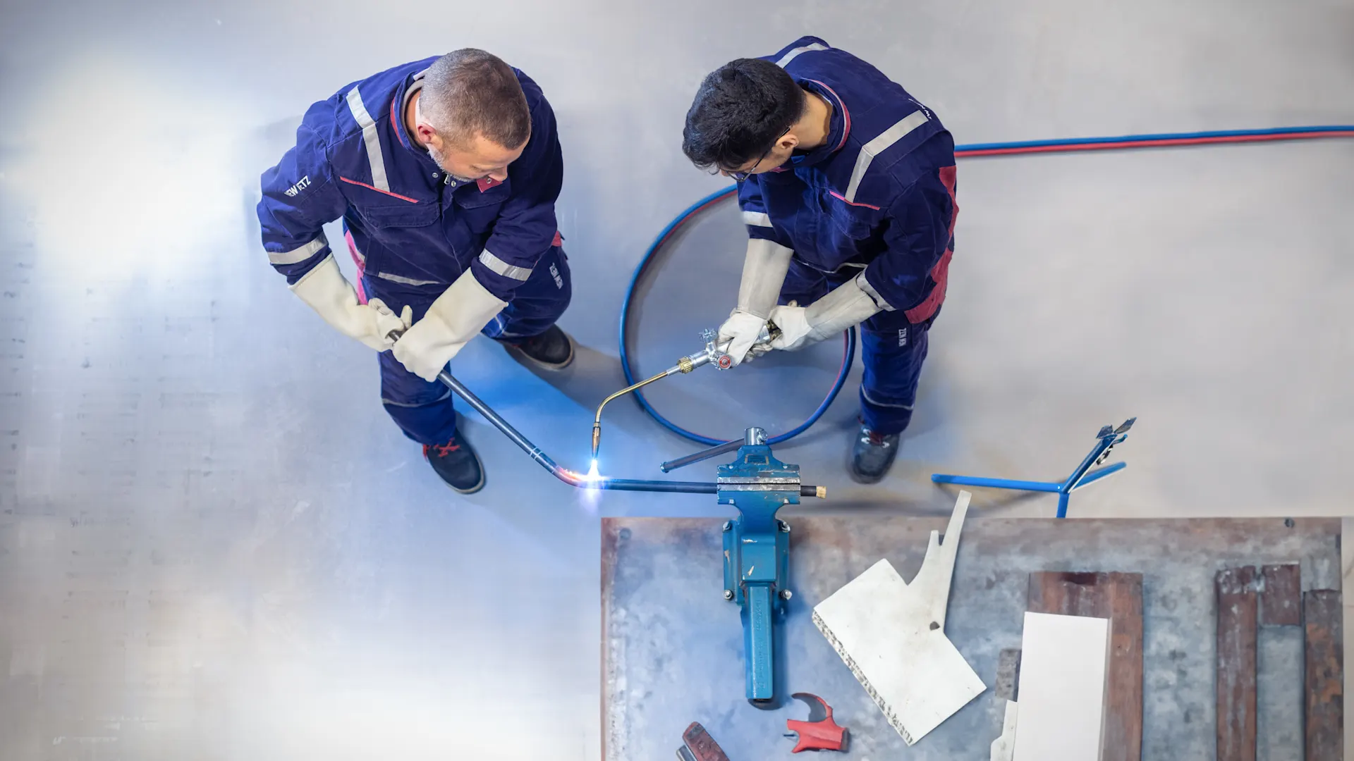 Wie Altersvielfalt Unternehmen stärkt Zwei Arbeiter in blauen Schutzanzügen und Handschuhen arbeiten von oben betrachtet an einem Metallwerkstück auf dem Boden. Einer hält einen Schneidbrenner, während daneben blaue Schläuche und Werkzeuge liegen.
