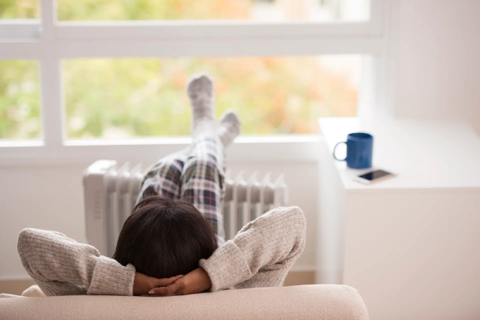 iStock-658574596 RuslanDashinsky Eine Person in karierter Pyjamahose und Wollsocken liegt entspannt auf einem beigen Sofa mit den Beinen nach oben an einer Heizung gelehnt. Im Hintergrund steht eine blaue Tasse auf einer Fensterbank vor einem großen Fenster.