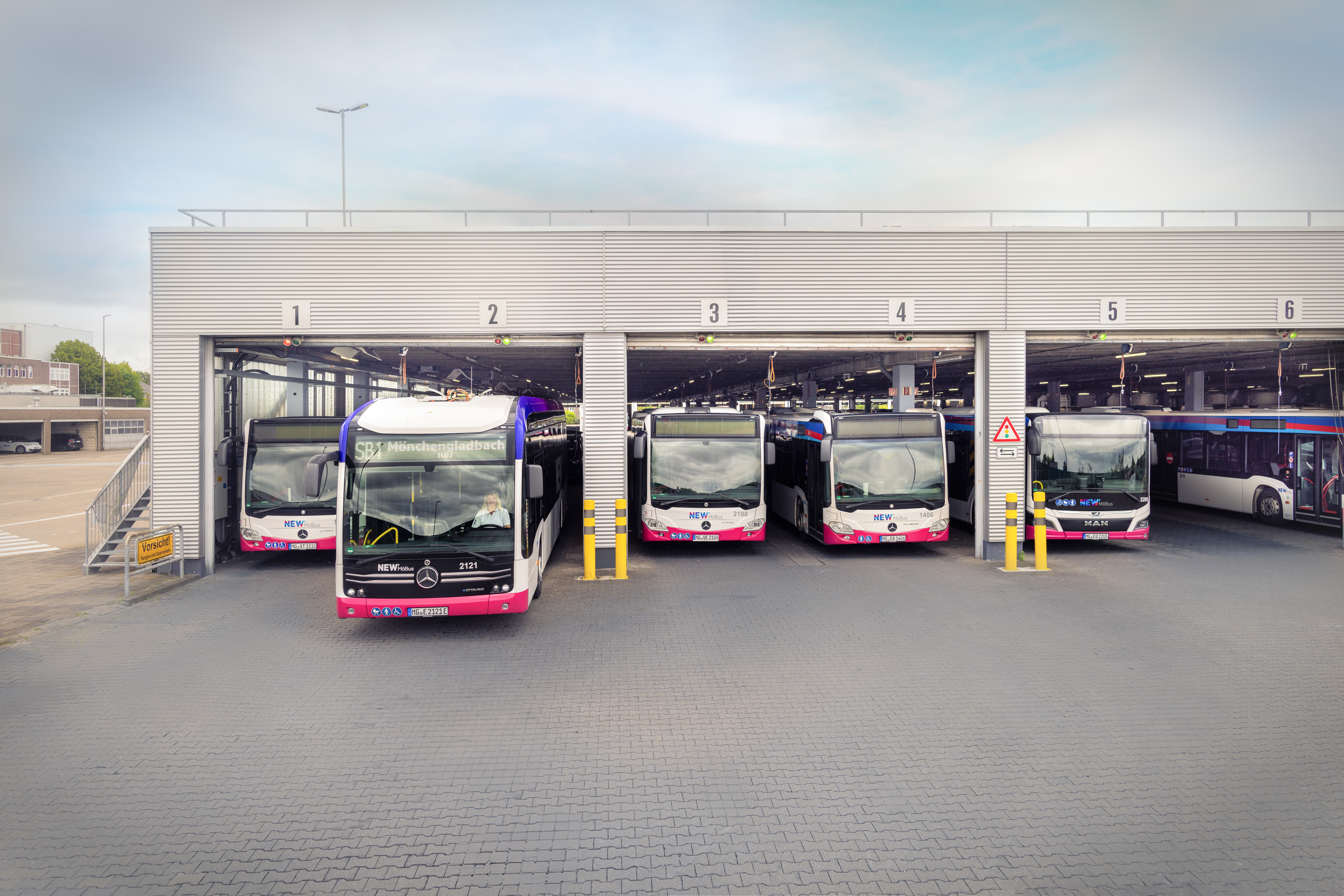 Mehrere Elektrobusse stehen in den Stellplätzen einer modernen Busgarage. Ein Bus fährt gerade aus der Halle heraus, die einzelnen Stellplätze sind nummeriert.