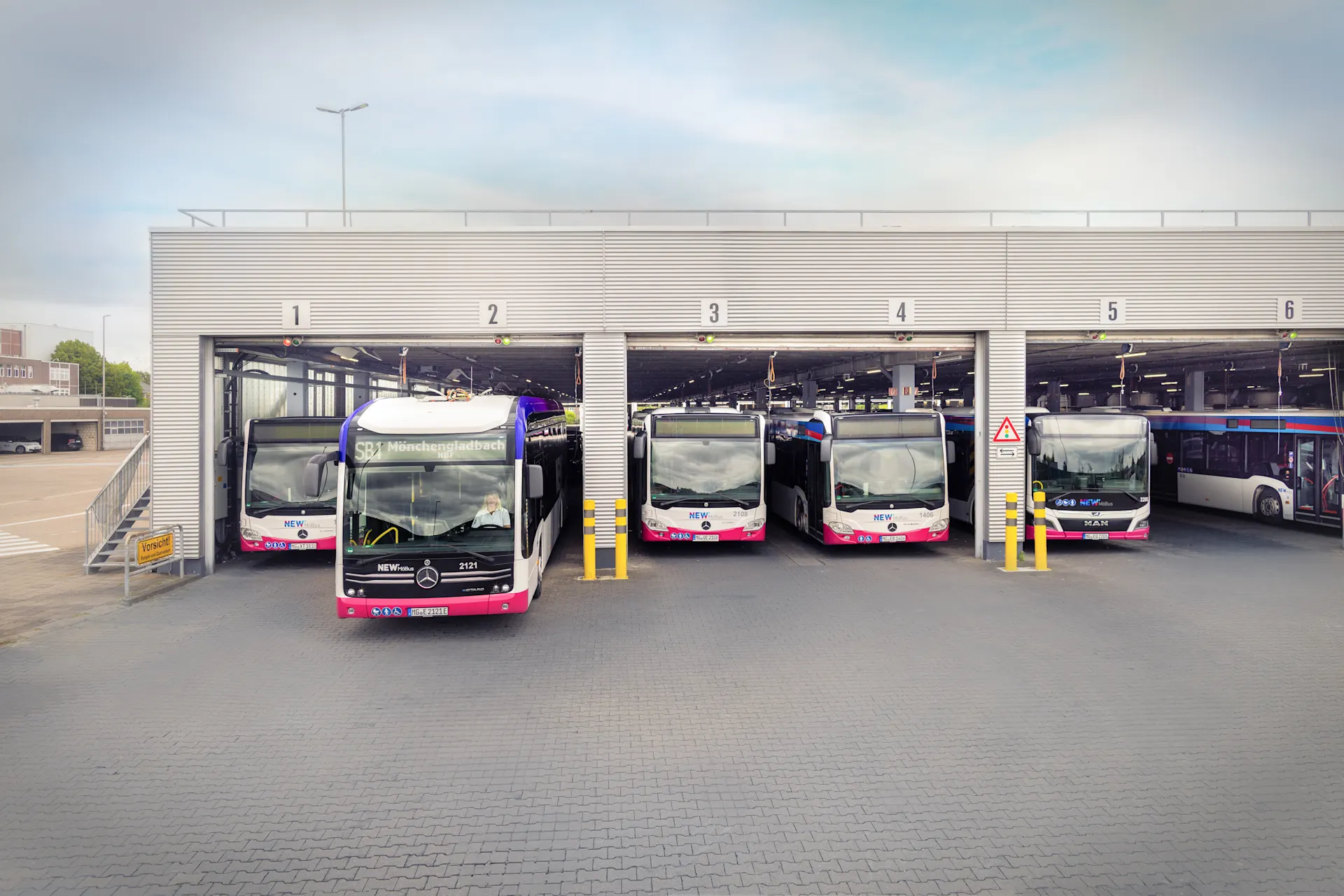 Mehrere Elektrobusse stehen in den Stellplätzen einer modernen Busgarage. Ein Bus fährt gerade aus der Halle heraus, die einzelnen Stellplätze sind nummeriert.
