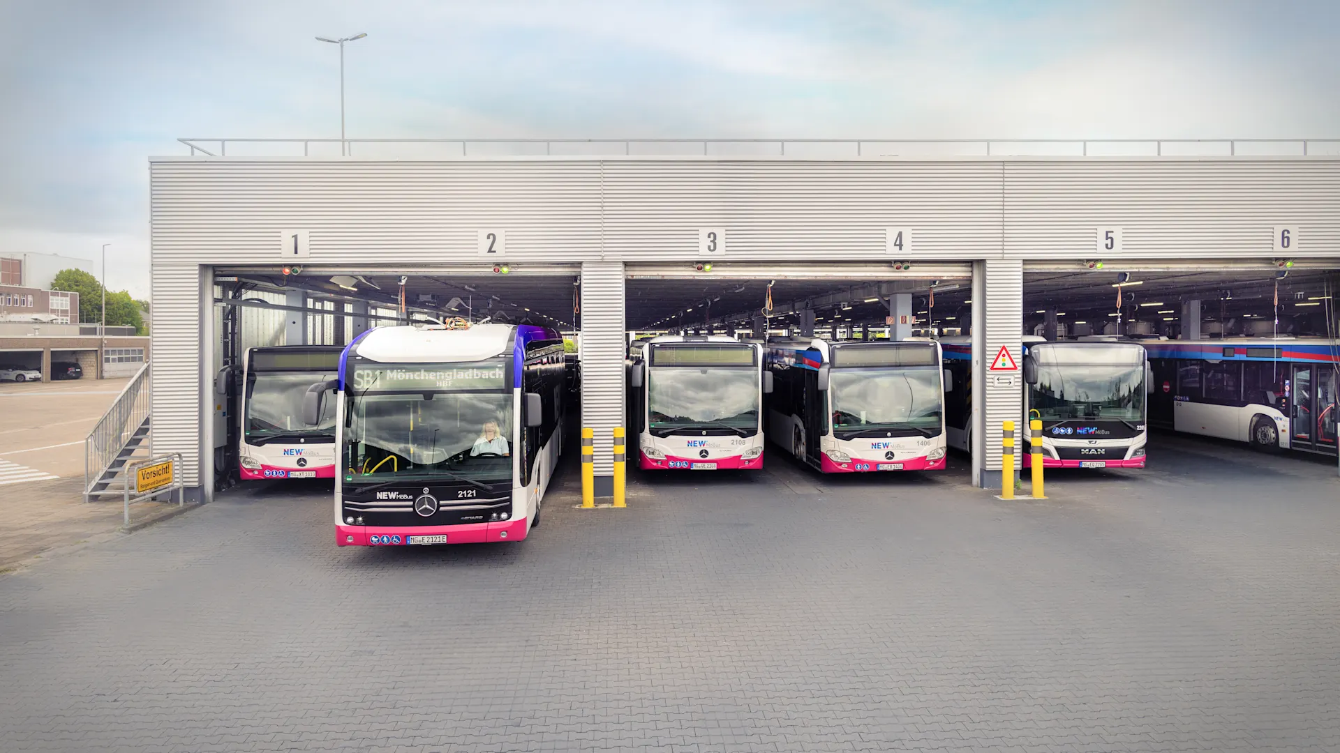 Clever unterwegs, nachhaltig voran. Mehrere Elektrobusse stehen in den Stellplätzen einer modernen Busgarage. Ein Bus fährt gerade aus der Halle heraus, die einzelnen Stellplätze sind nummeriert.