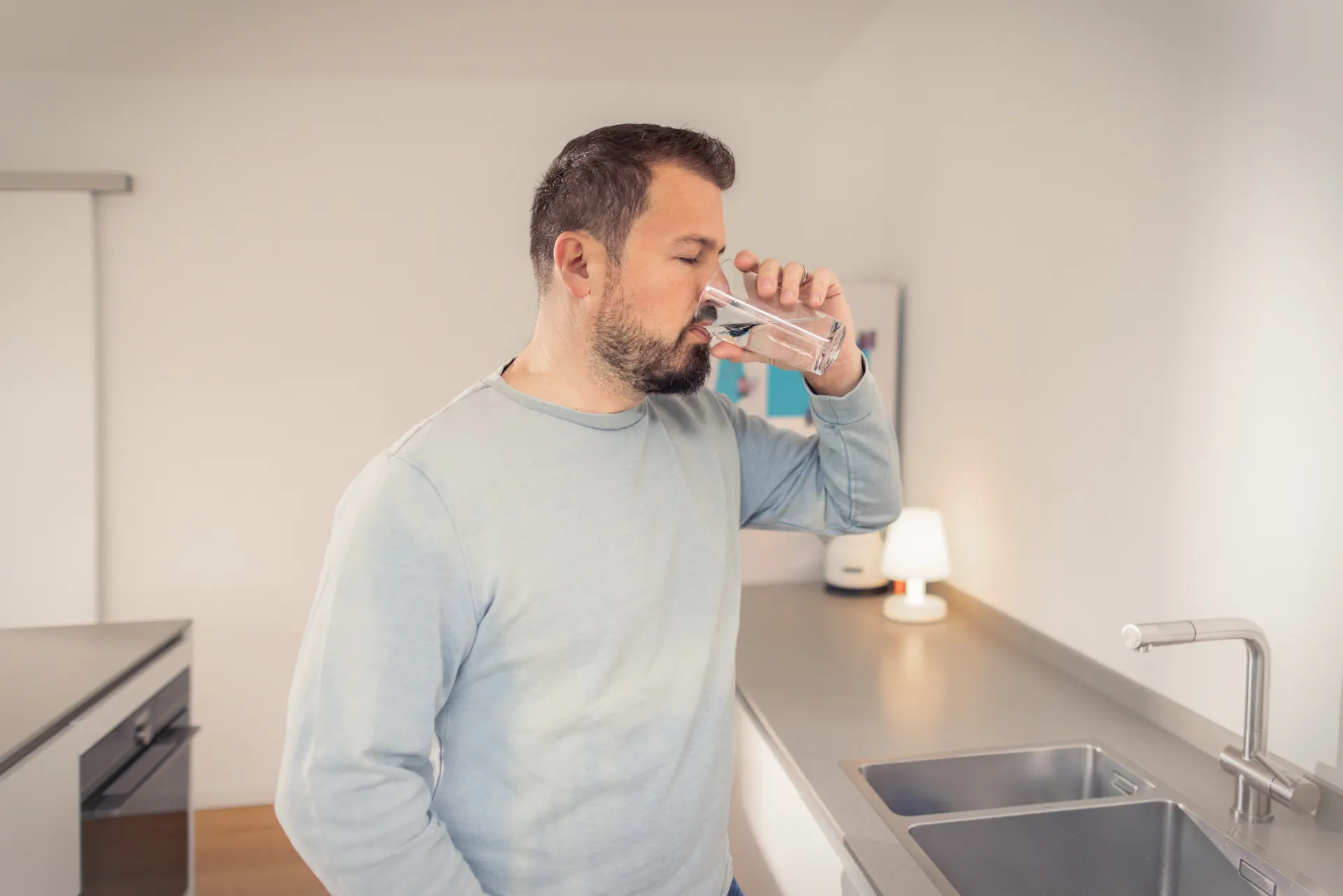 Trinkwasser - Lassen Sie Ihrem Durst freien Lauf Ein Mann steht in der Küche und trinkt Leitungswasser aus einem Glas