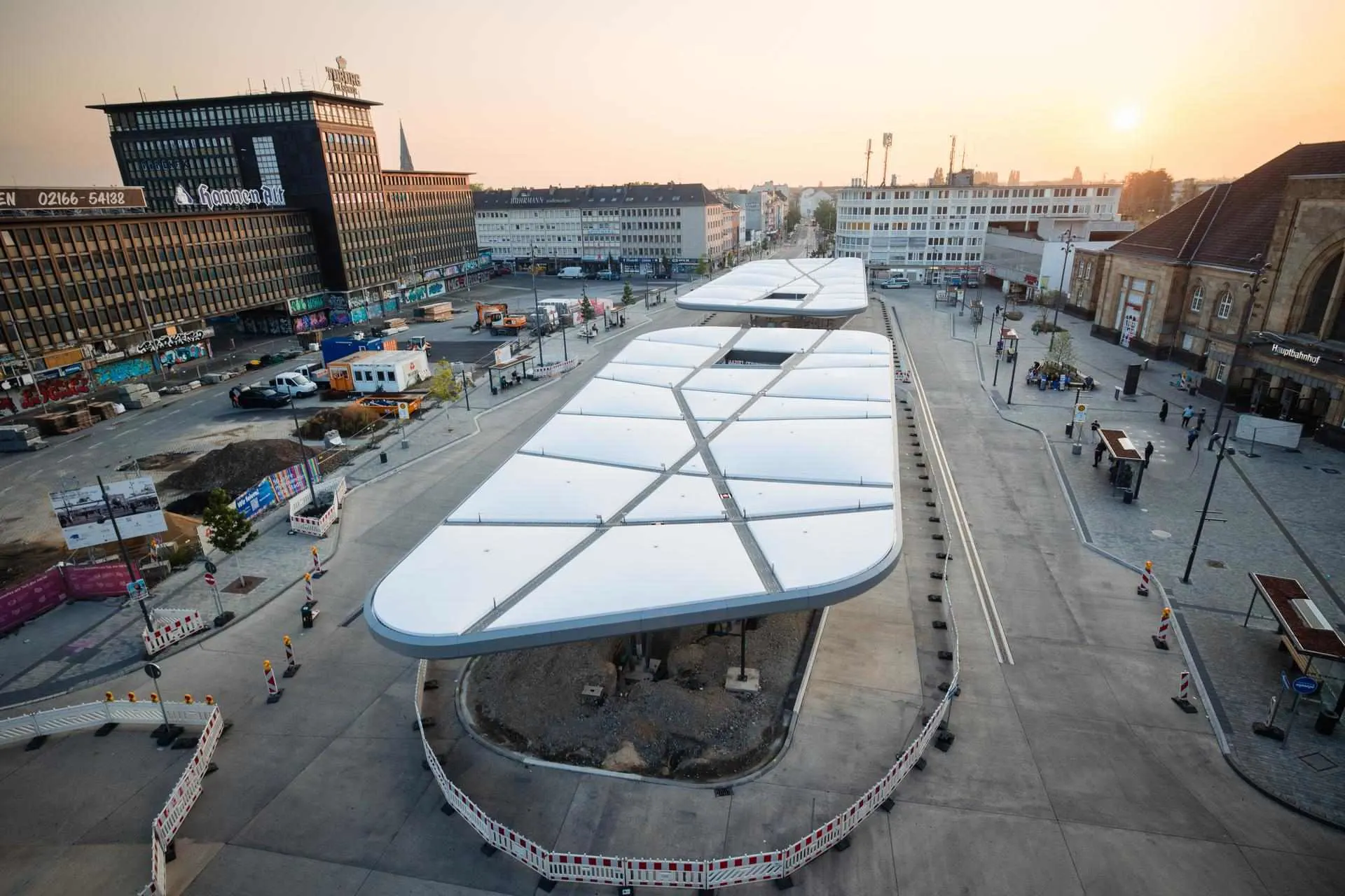 Baum-4725 Das Bild zeigt den Zentralen Omnibusbahnhof (ZOB) in Mönchengladbach bei Sonnenaufgang. Die moderne Dachkonstruktion prägt das Stadtbild. Rechts im Bild befindet sich der Hauptbahnhof, links das markante Gebäude „Haus Westland“.