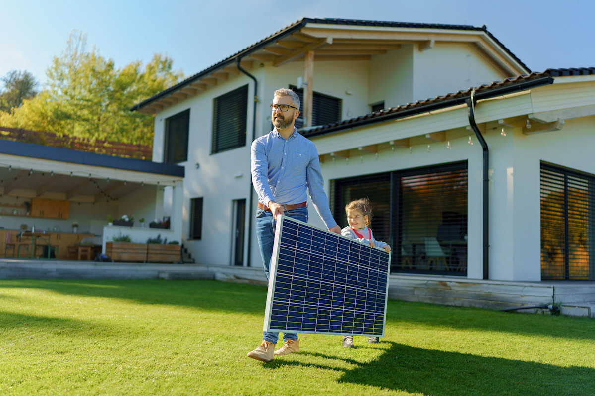 Ein Mann in blauem Hemd hält zusammen mit einem Kind ein Solarmodul in den Händen und läuft damit über eine Wiese. Das Haus hinter ihnen hat eine helle Fassade und Fenster die große Teile der Fassade einnehmen.