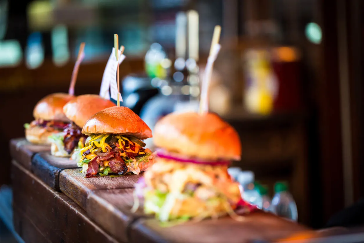 Street Food Festival (Foto: iStock / coldsnowstorm) Nahaufnahme einer Auswahl frisch gegrillter Burger in Reihe auf einer Holztheke, Jeder der Burger hat sein eigenes Etikett, auf dem der Inhalt des Burgers steht. Die Burger werden zwischen glasierten Brötchen eingeklemmt und auf Betten mit frischem grünem Salat präsentiert und mit geschmolzenem Käse und roten Zwiebeln gefüllt. (Copyright coldsnowstorm Stock-Fotografie-ID:914886270)